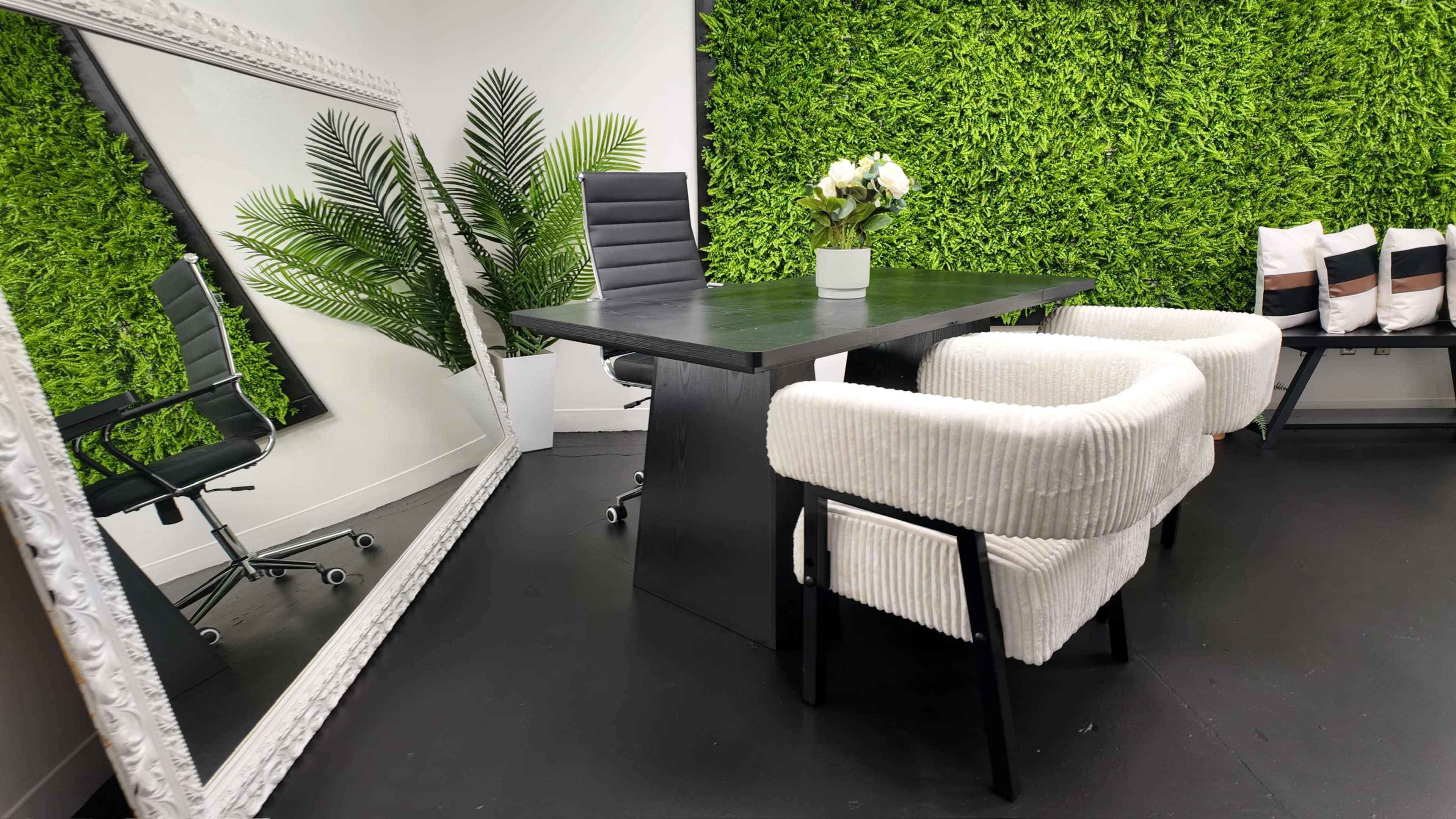 The scene features a modern office space with a black table and two white fabric chairs, a large mirror, and a wall adorned with green foliage.