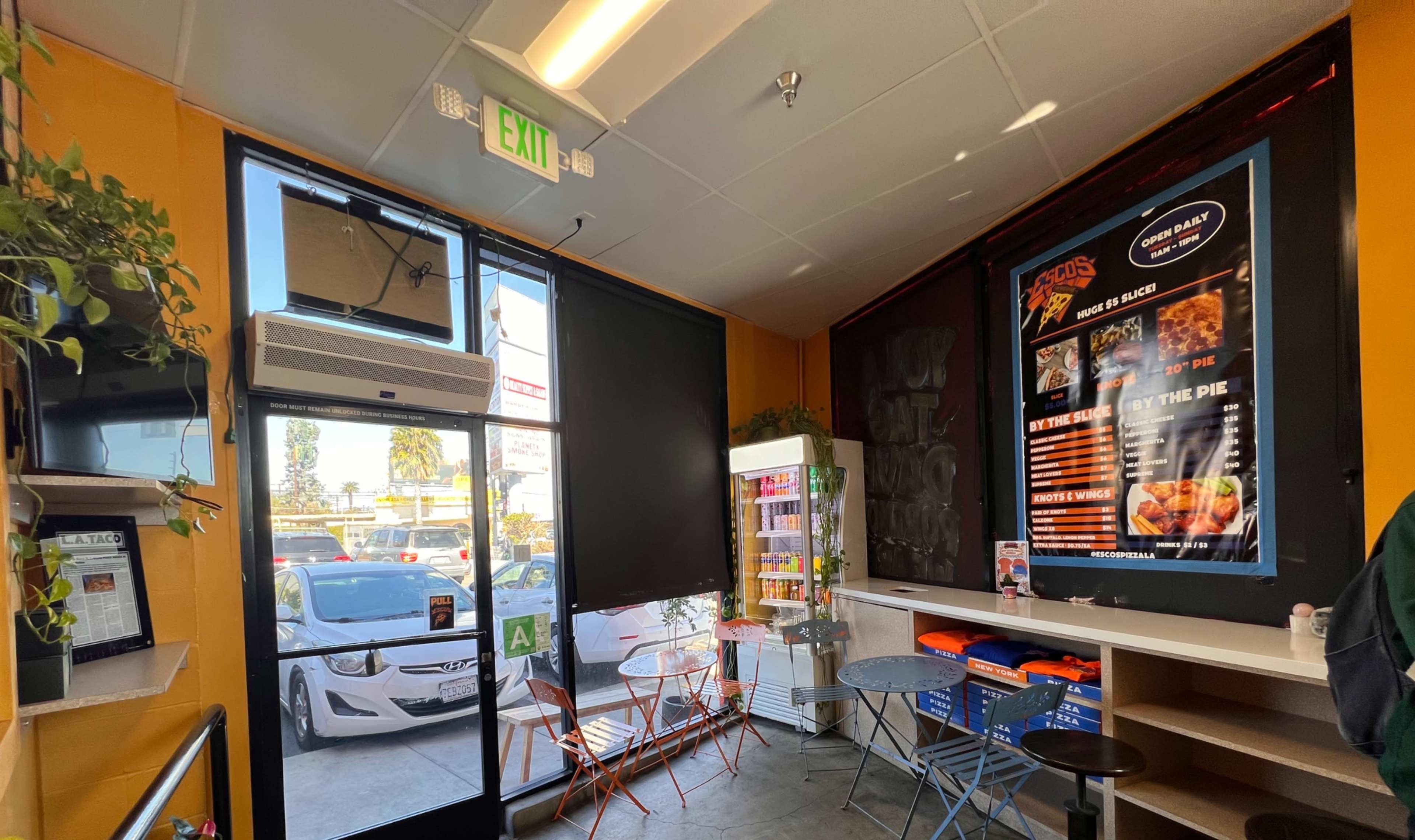 The image shows the interior of a small eatery with colorful chairs and tables, a menu displayed on a wall, and a view of parked cars outside through a large window.