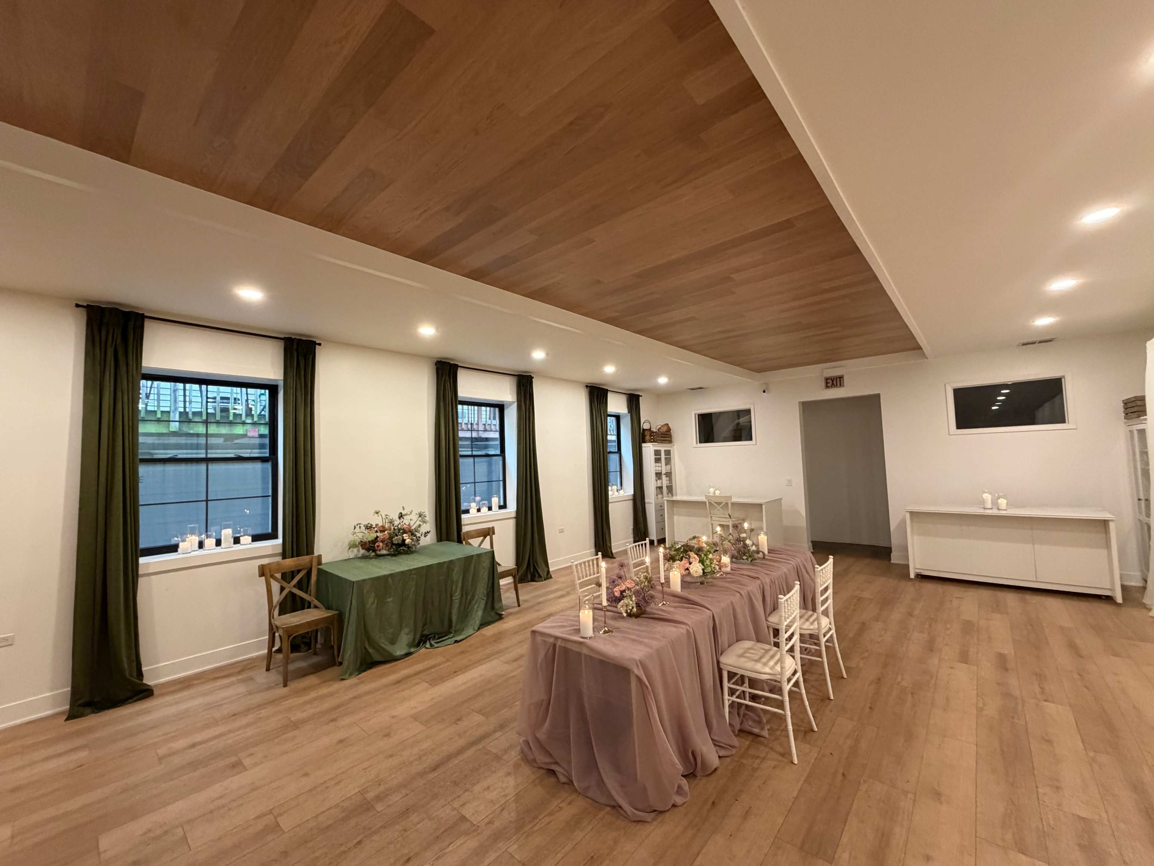 The image shows a spacious indoor room with wooden flooring, featuring a long table set with light-colored linens and decorations, flanked by white chairs and large windows with green curtains.
