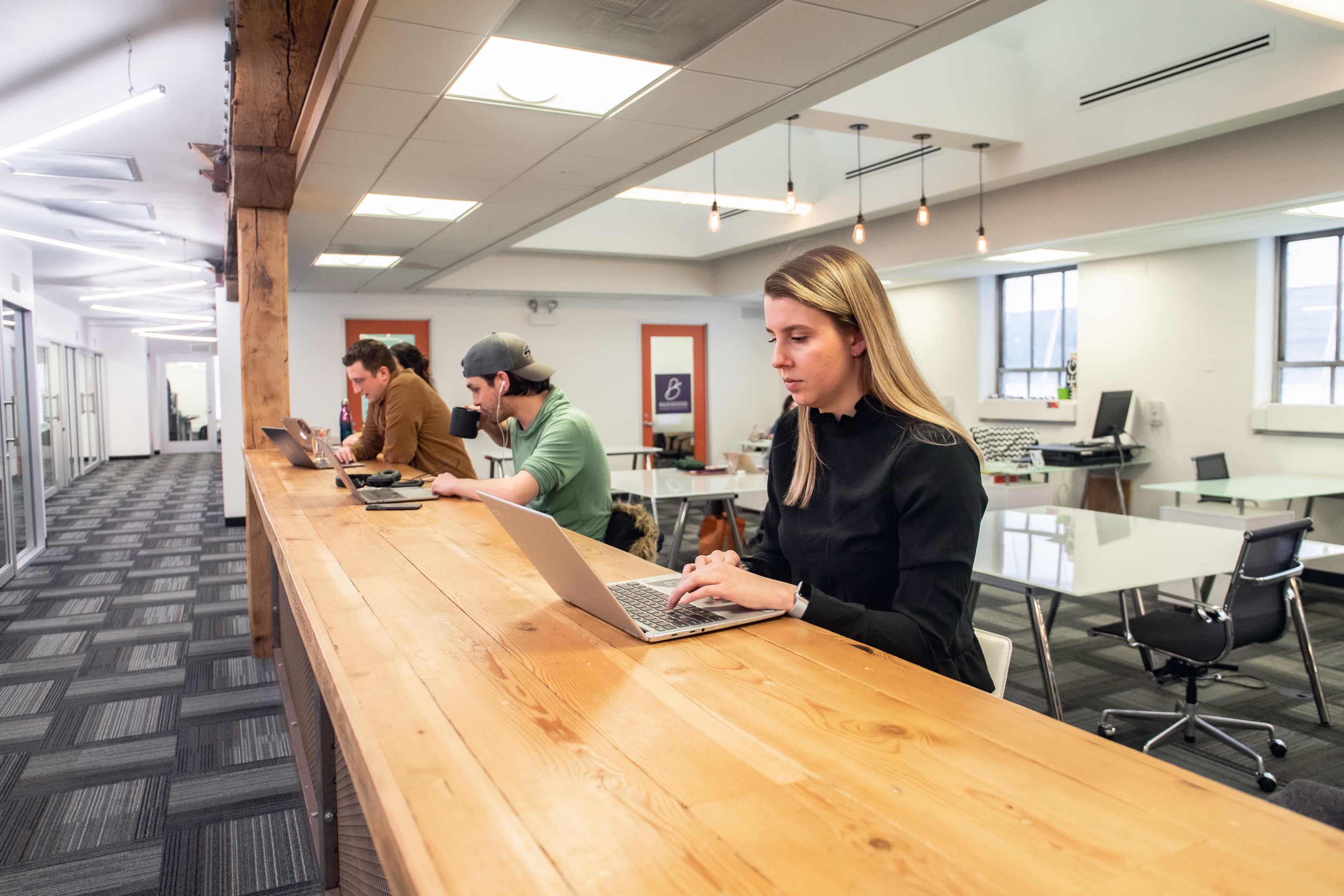 A group of four individuals works on laptops at a long wooden counter in a modern office space.