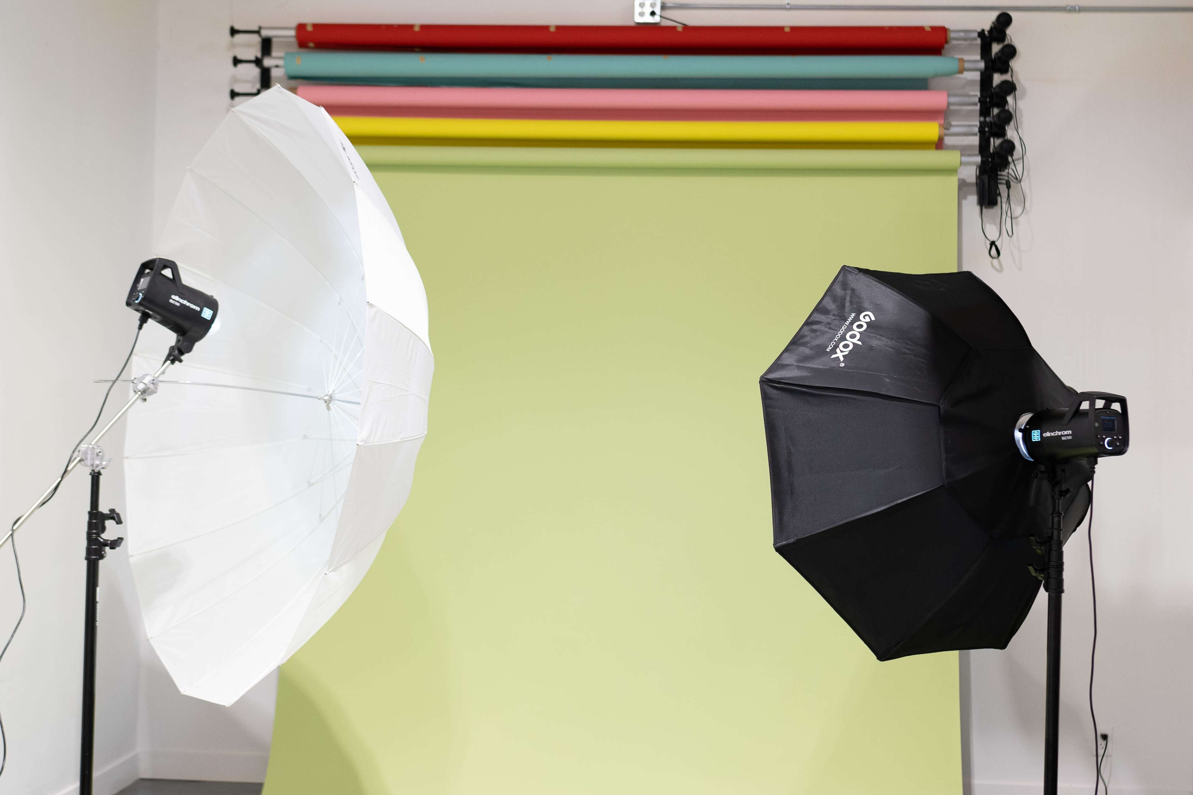 Two studio light umbrellas, one white and one black, positioned in front of a green backdrop with colorful stripes mounted above it.