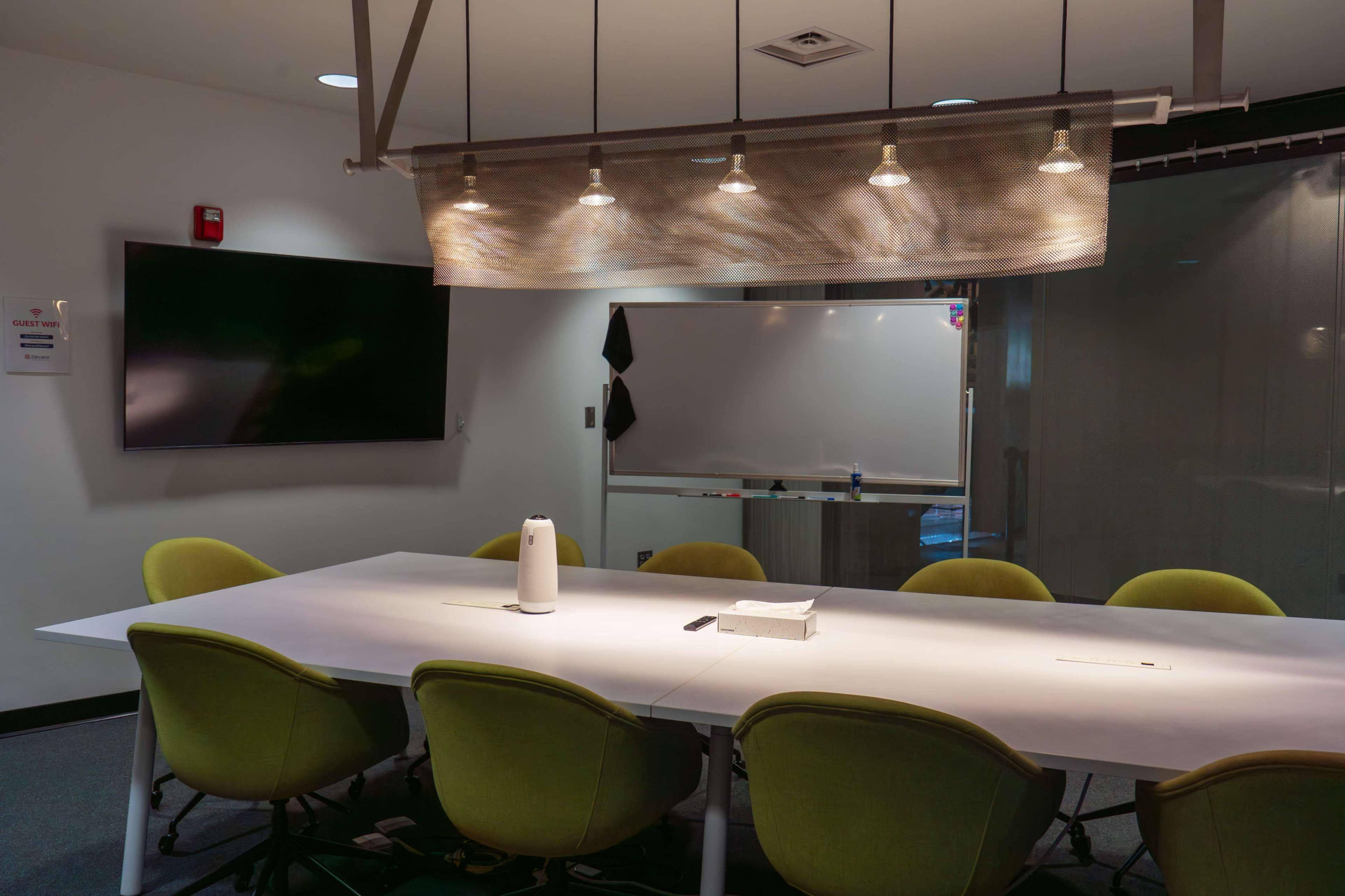 The image shows a modern conference room with a long white table surrounded by green chairs, a large screen on the wall, and a whiteboard in the background.