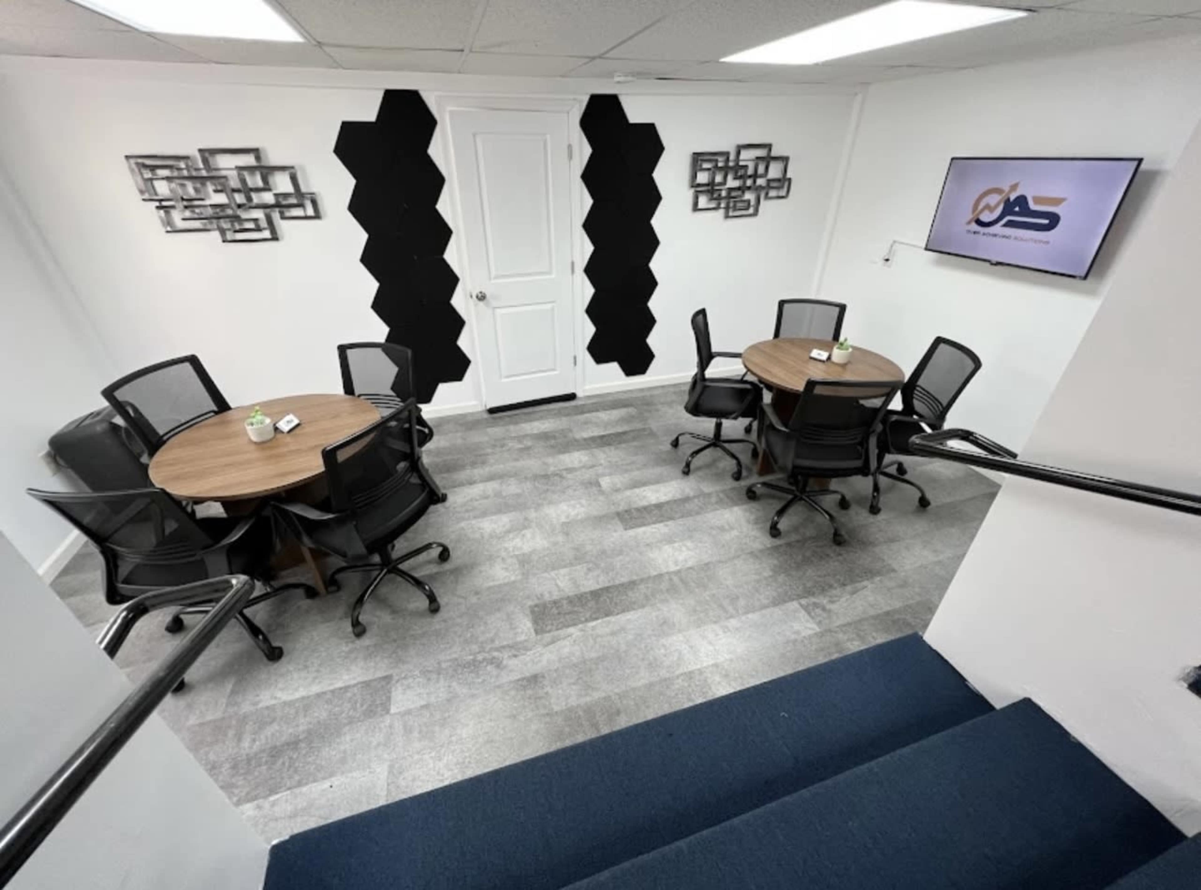 A modern office space with two round wooden tables surrounded by black chairs and a TV mounted on the wall.