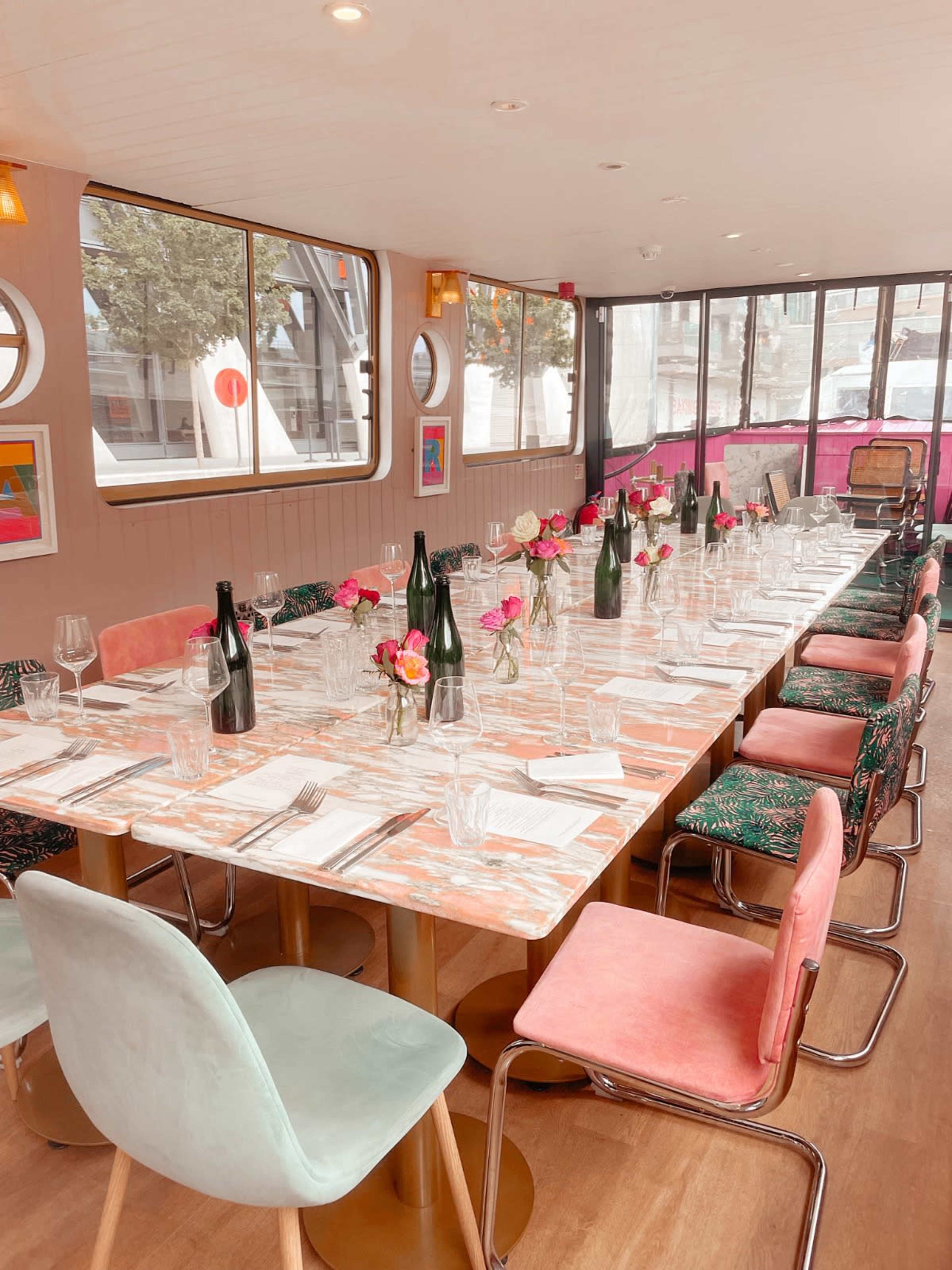 A long dining table is set with glassware and flower centerpieces in a brightly lit room featuring large windows and colorful decor.