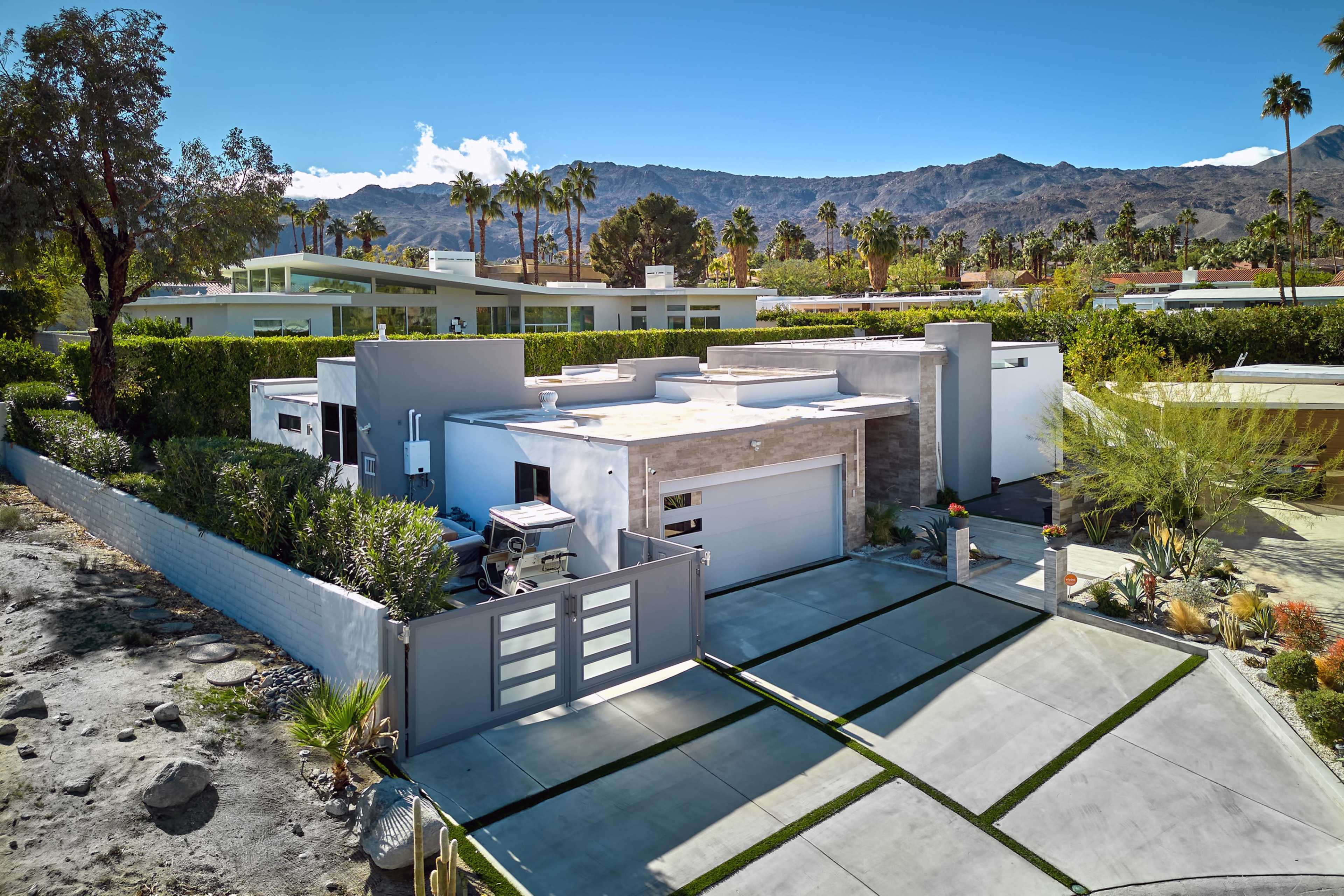 A modern one-story house with a garage and a landscaped driveway, set against a backdrop of mountains and palm trees.