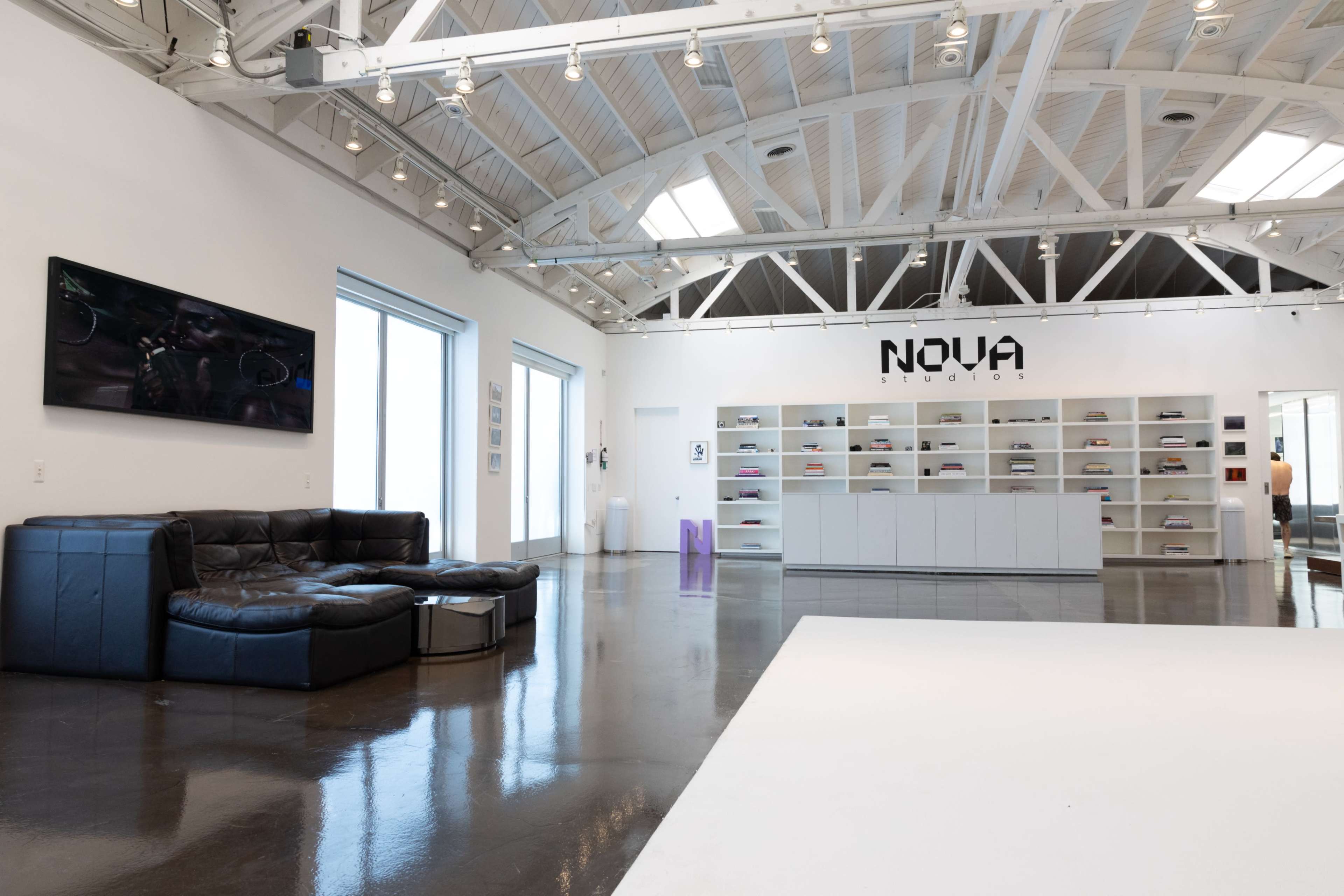 The image shows a spacious studio environment with high ceilings, featuring a black leather sofa, a wall-mounted television, and a shelving unit displaying various items, with "NOVA" prominently displayed on the wall.