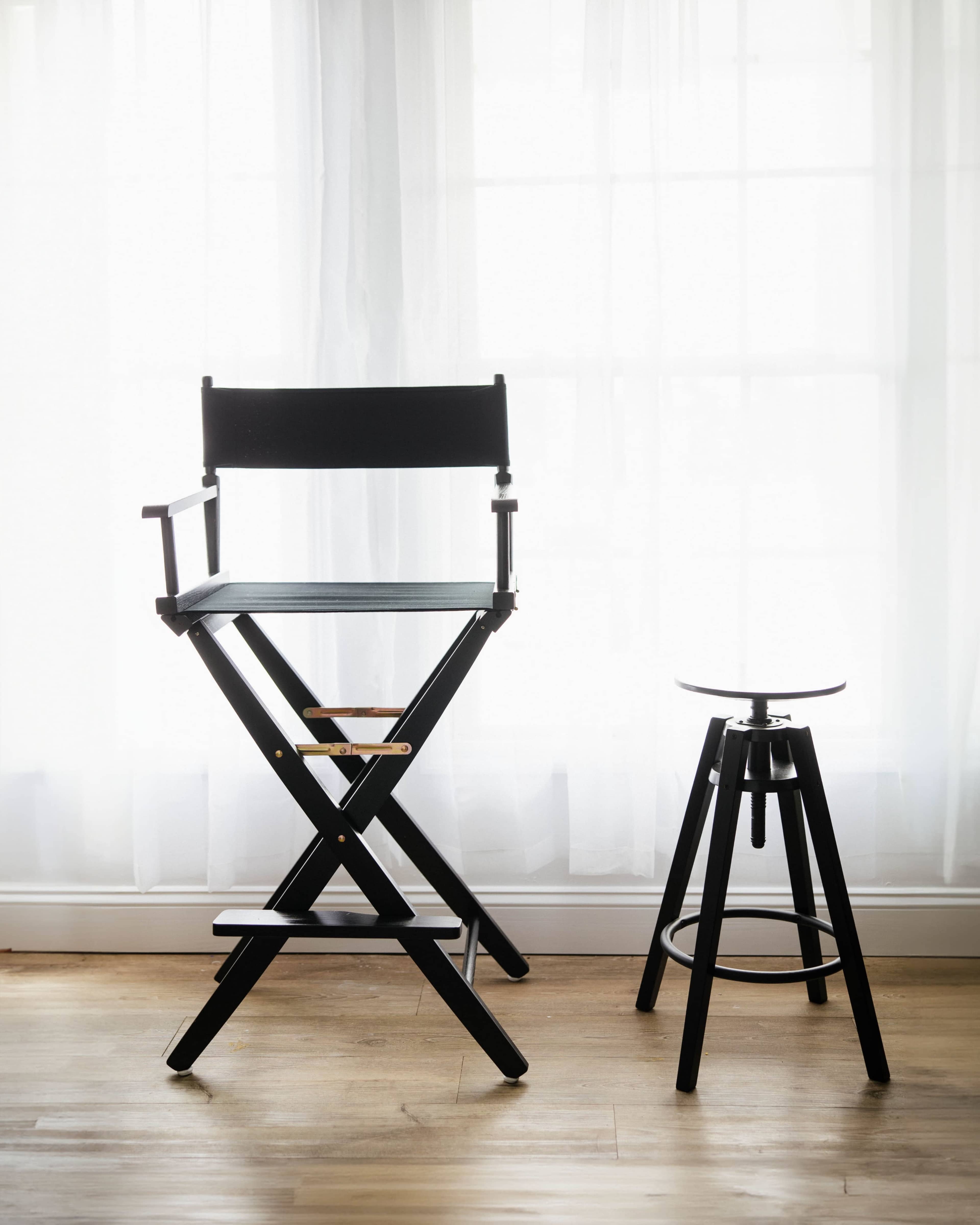 A black director's chair and a black stool stand in front of a window with sheer white curtains.