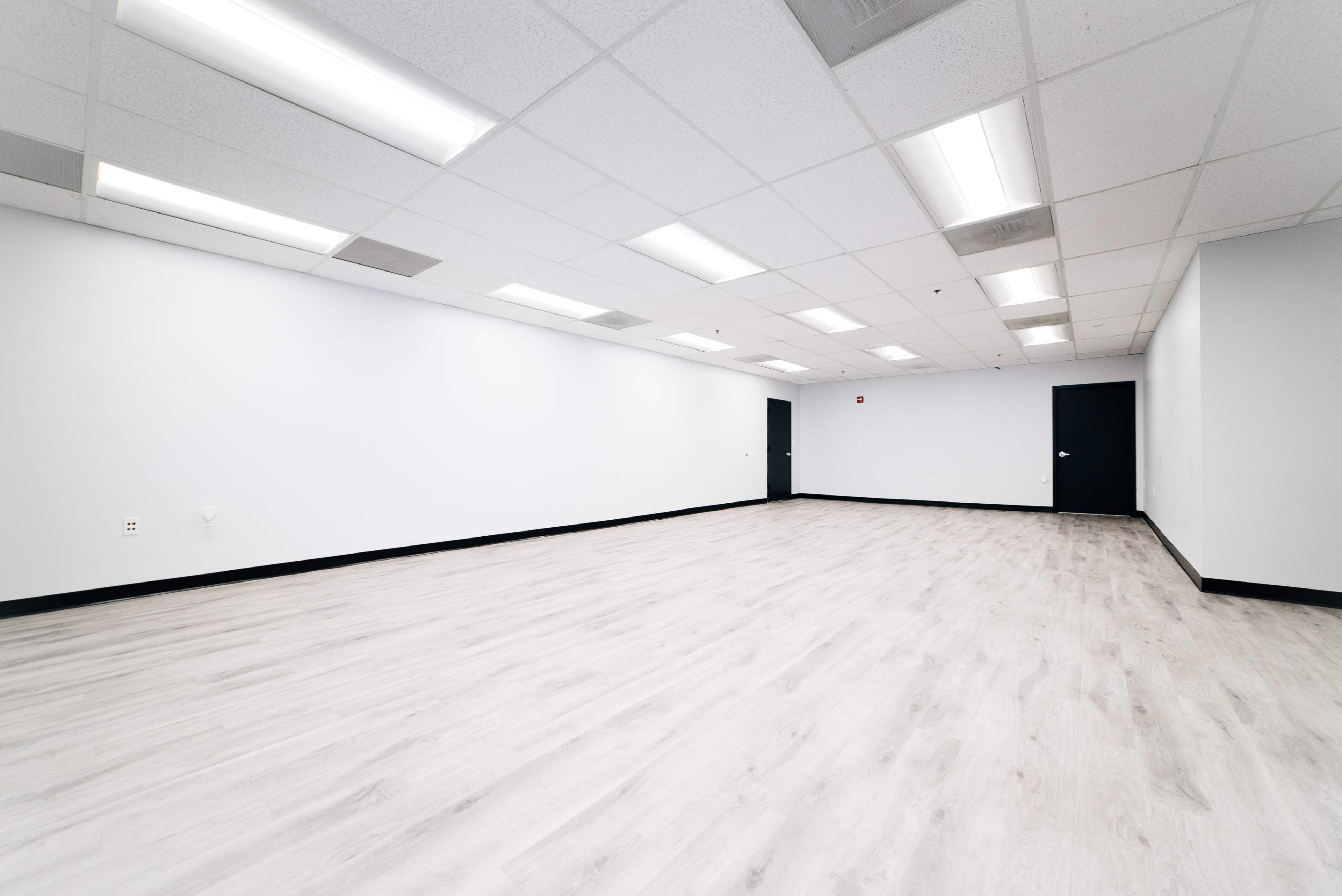 The image shows a spacious empty room with light-colored flooring, white walls, and fluorescent ceiling lights.