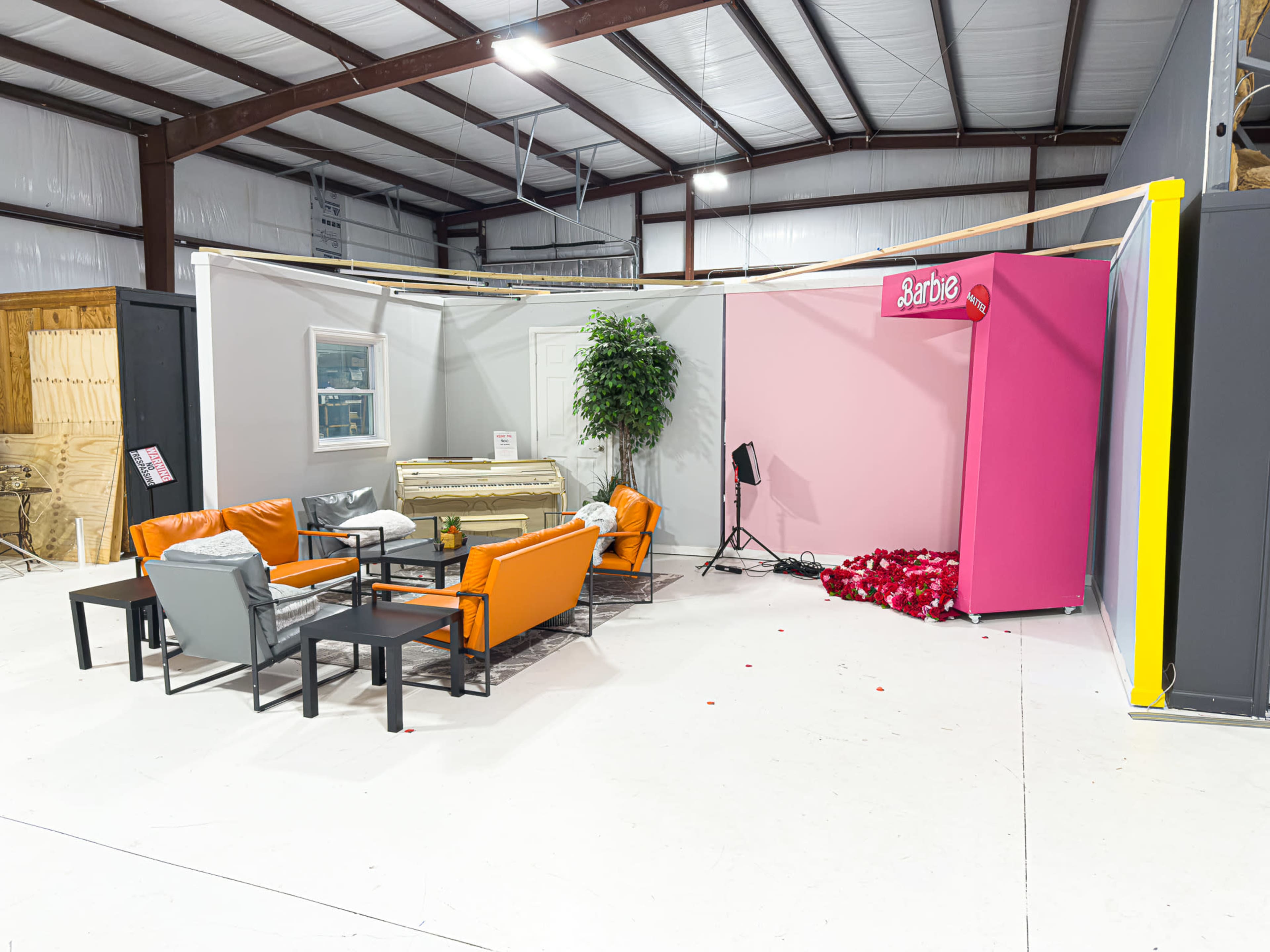 The image shows a staged interior space featuring a seating area with orange and gray sofas, a decorative piano, and a large pink display labeled 'Barbie'.
