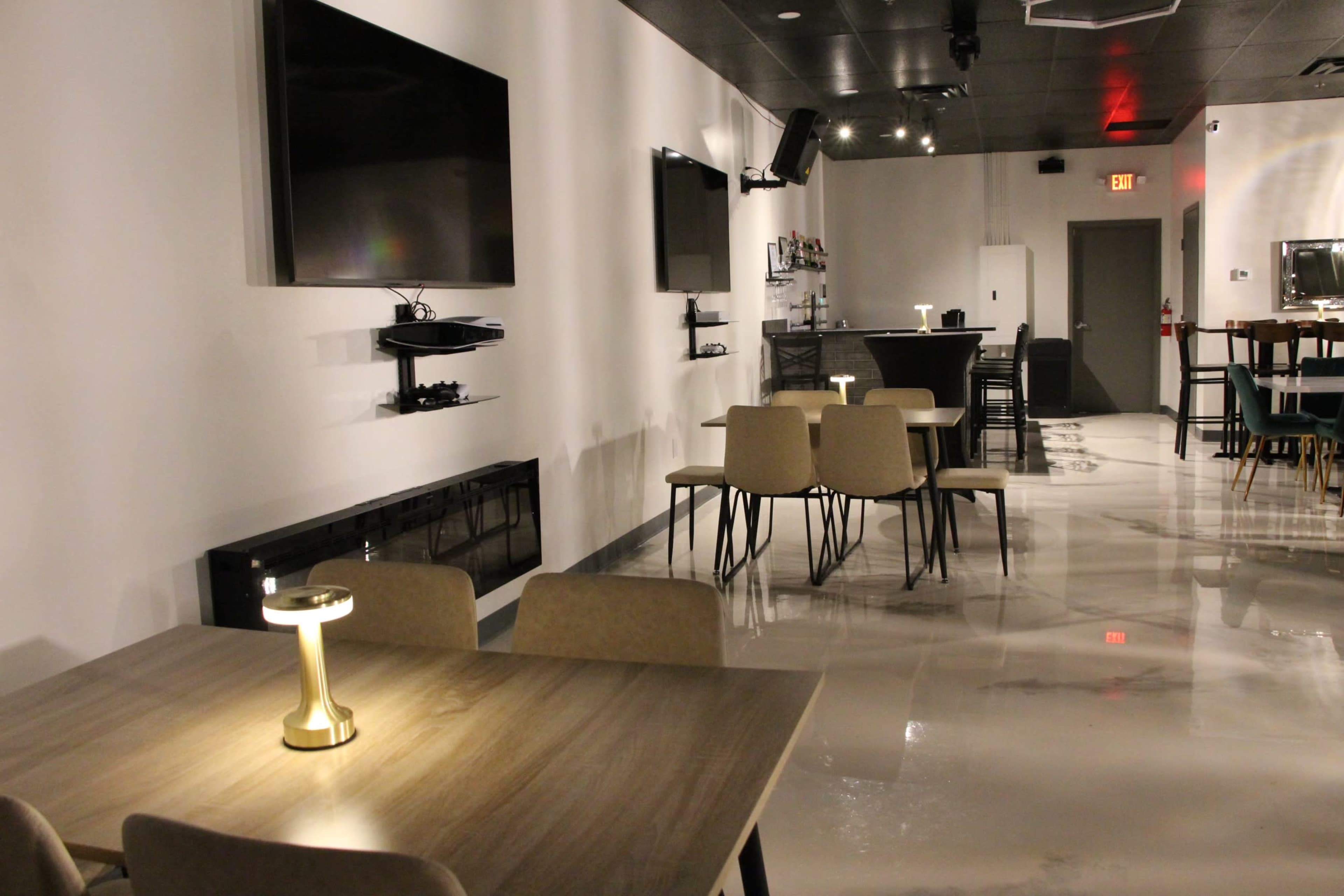 A modern lounge area with several tables and chairs, wall-mounted TVs, and a bar in the background.