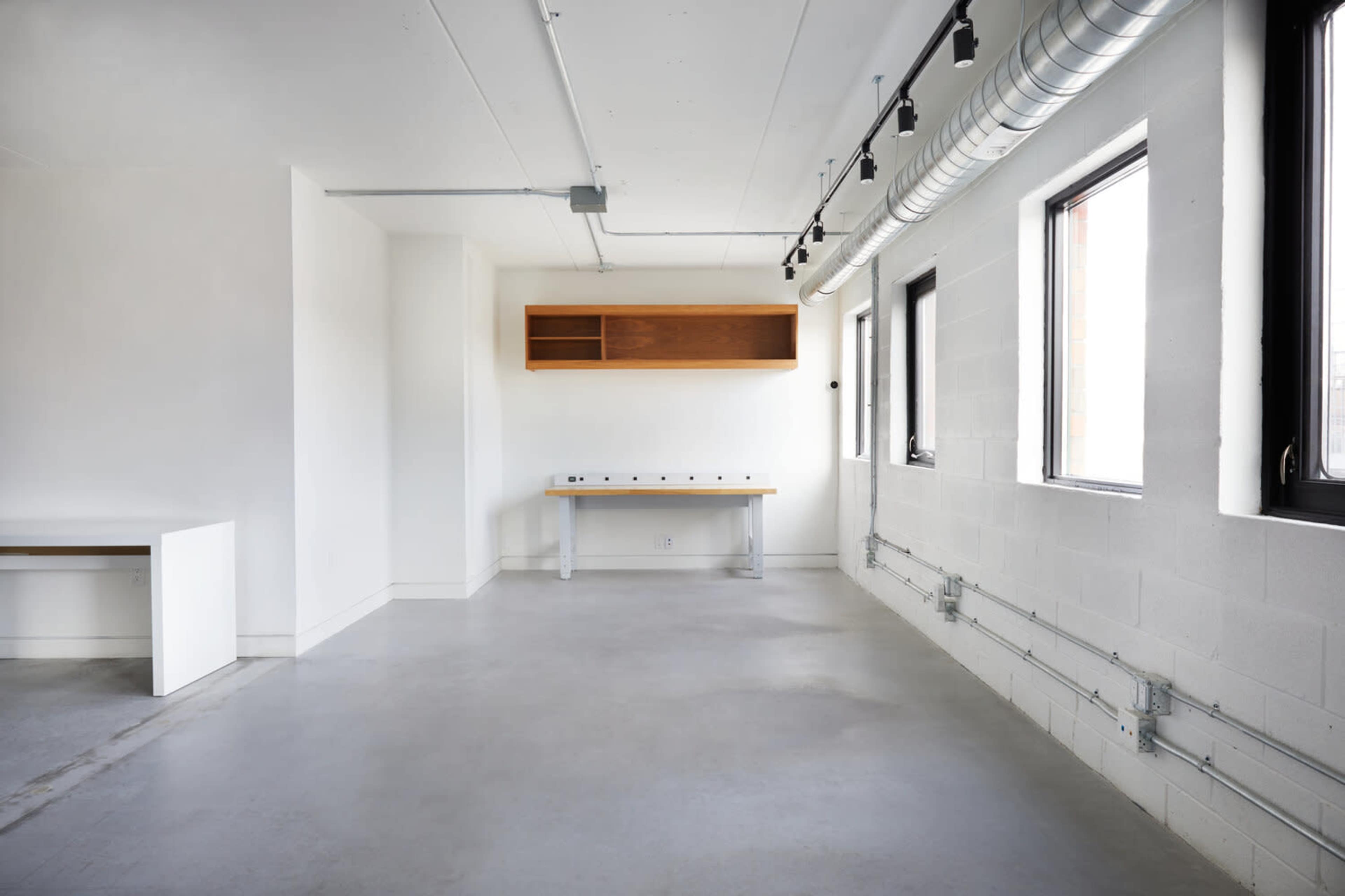 The image shows a spacious, empty room with concrete flooring, white walls, windows, and a wooden shelf mounted above a long table.