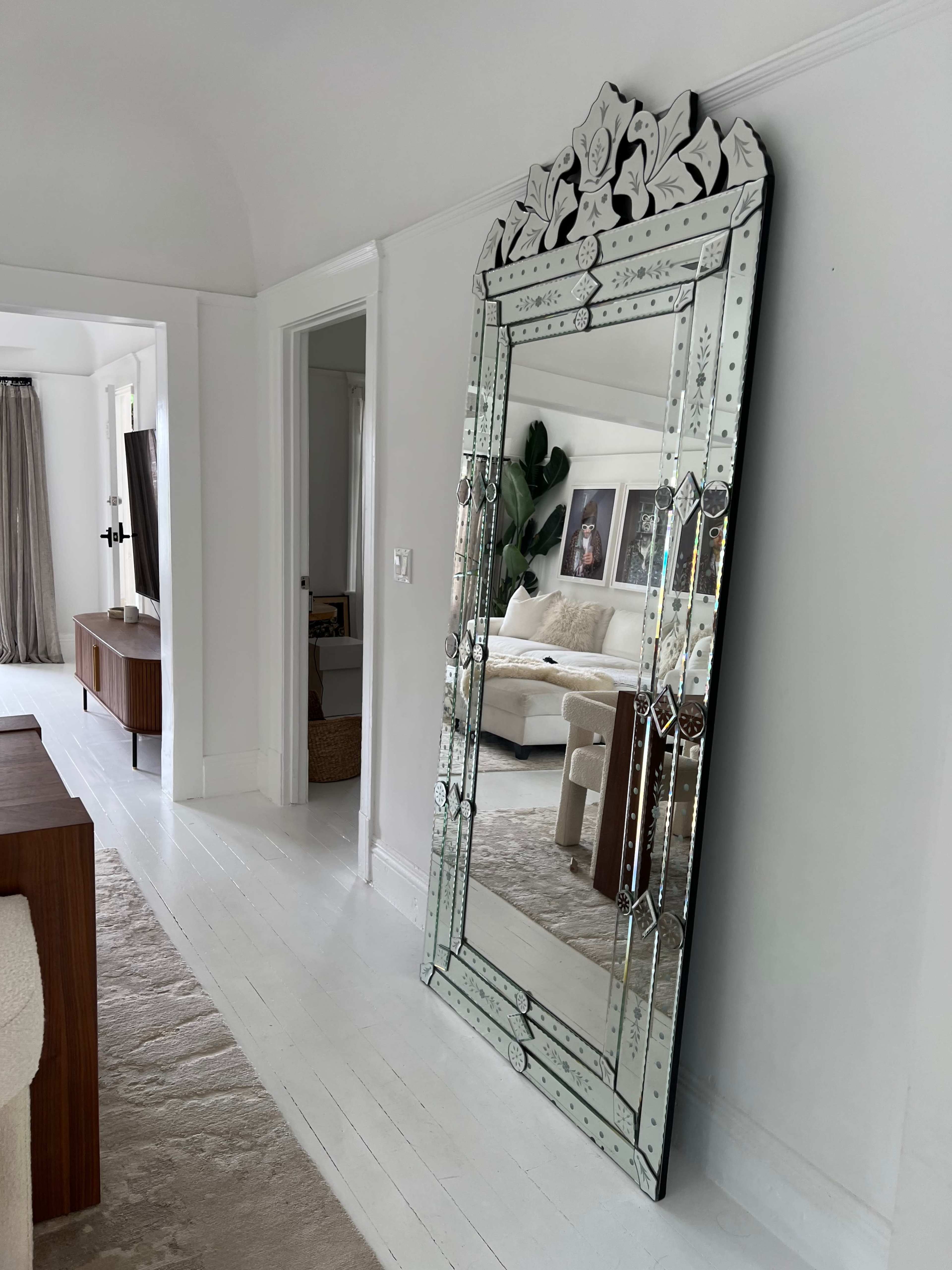 A large, ornate mirror stands against a white wall in a brightly lit hallway, reflecting the adjacent room.