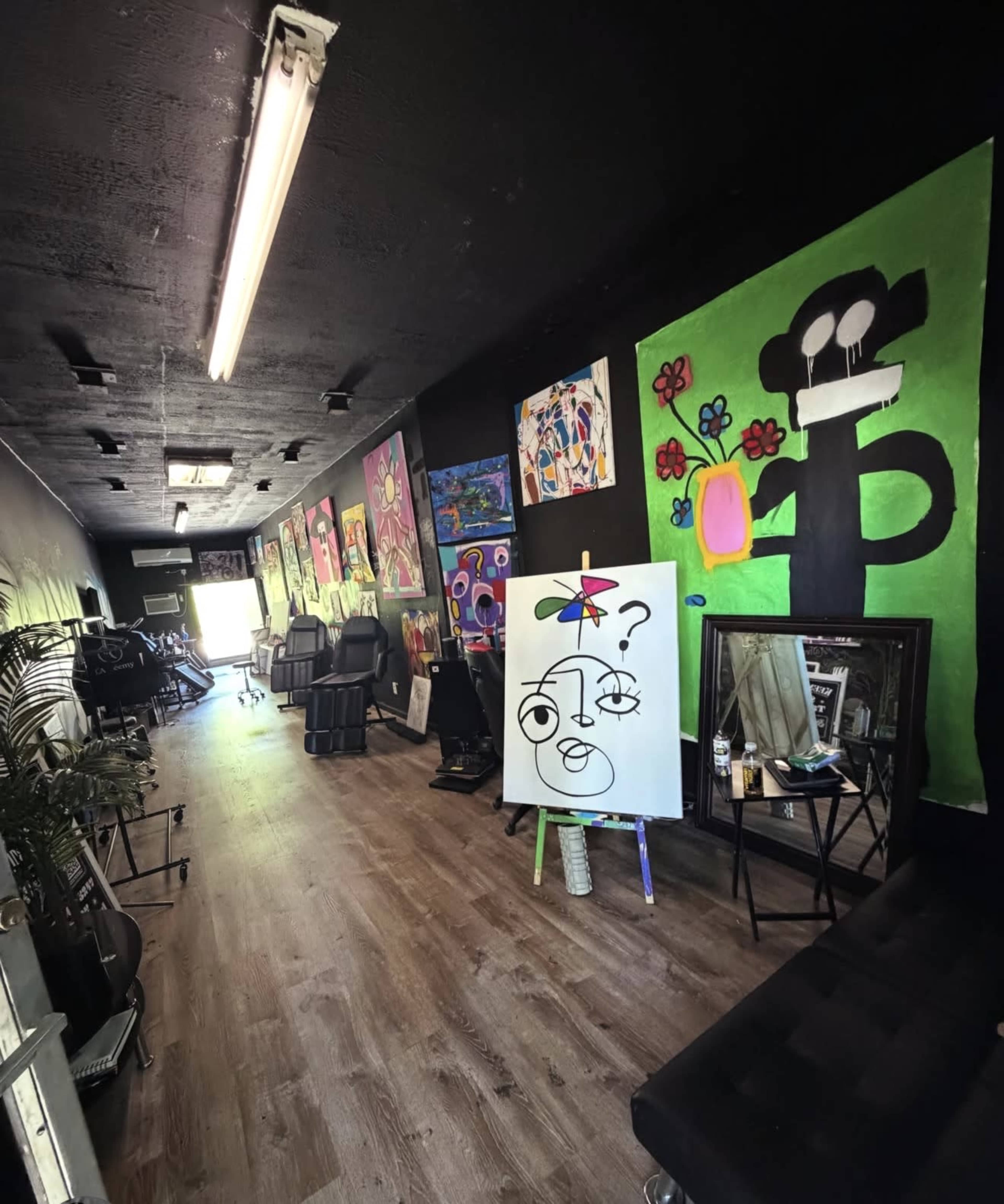 The image shows an art studio or gallery with colorful paintings displayed on the walls, featuring a large white canvas with a face sketch in the center.