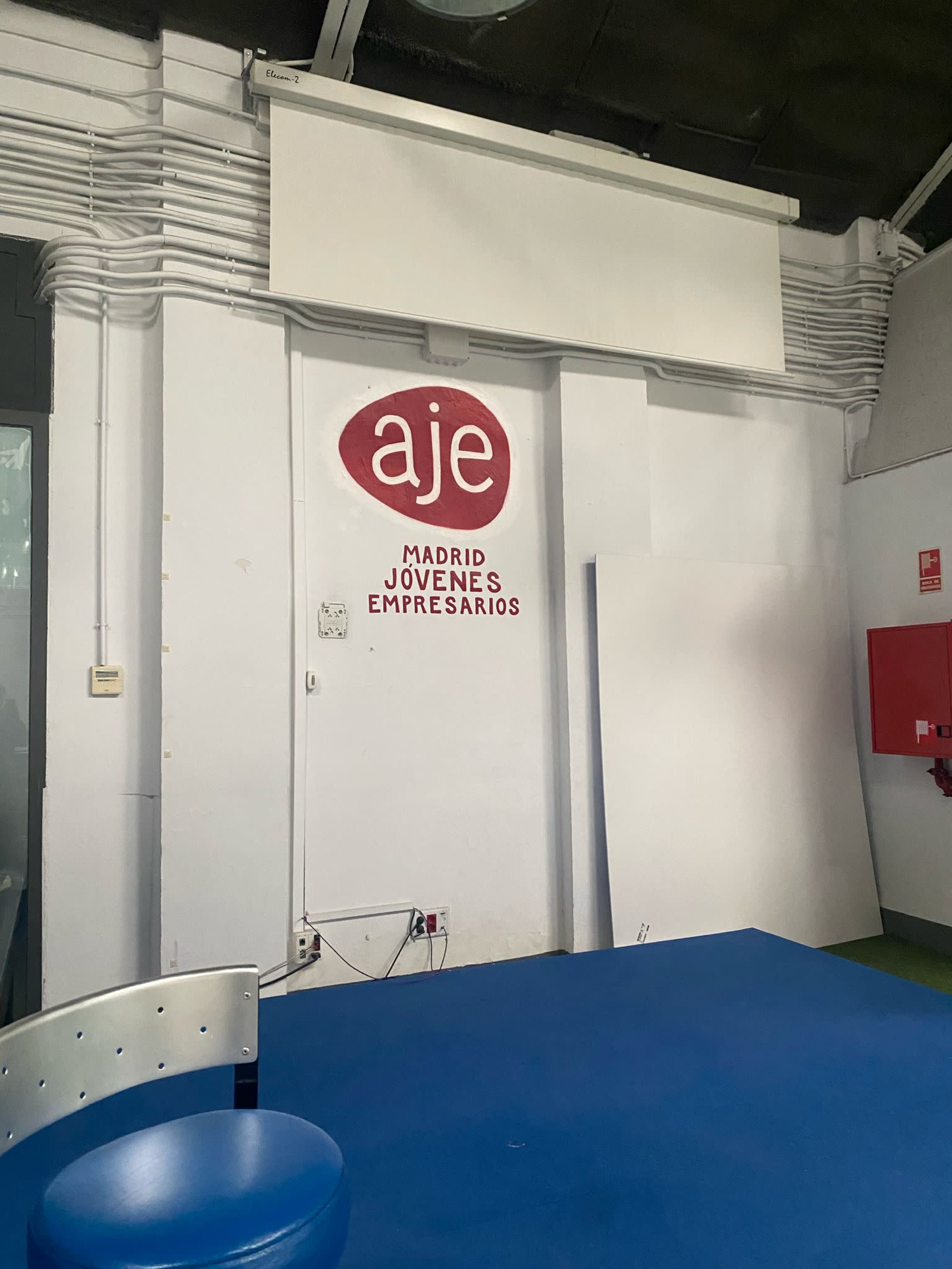 The image shows a wall in a room with a sign that reads "aje MADRID JÓVENES EMPRESARIOS" and a blue table below it.