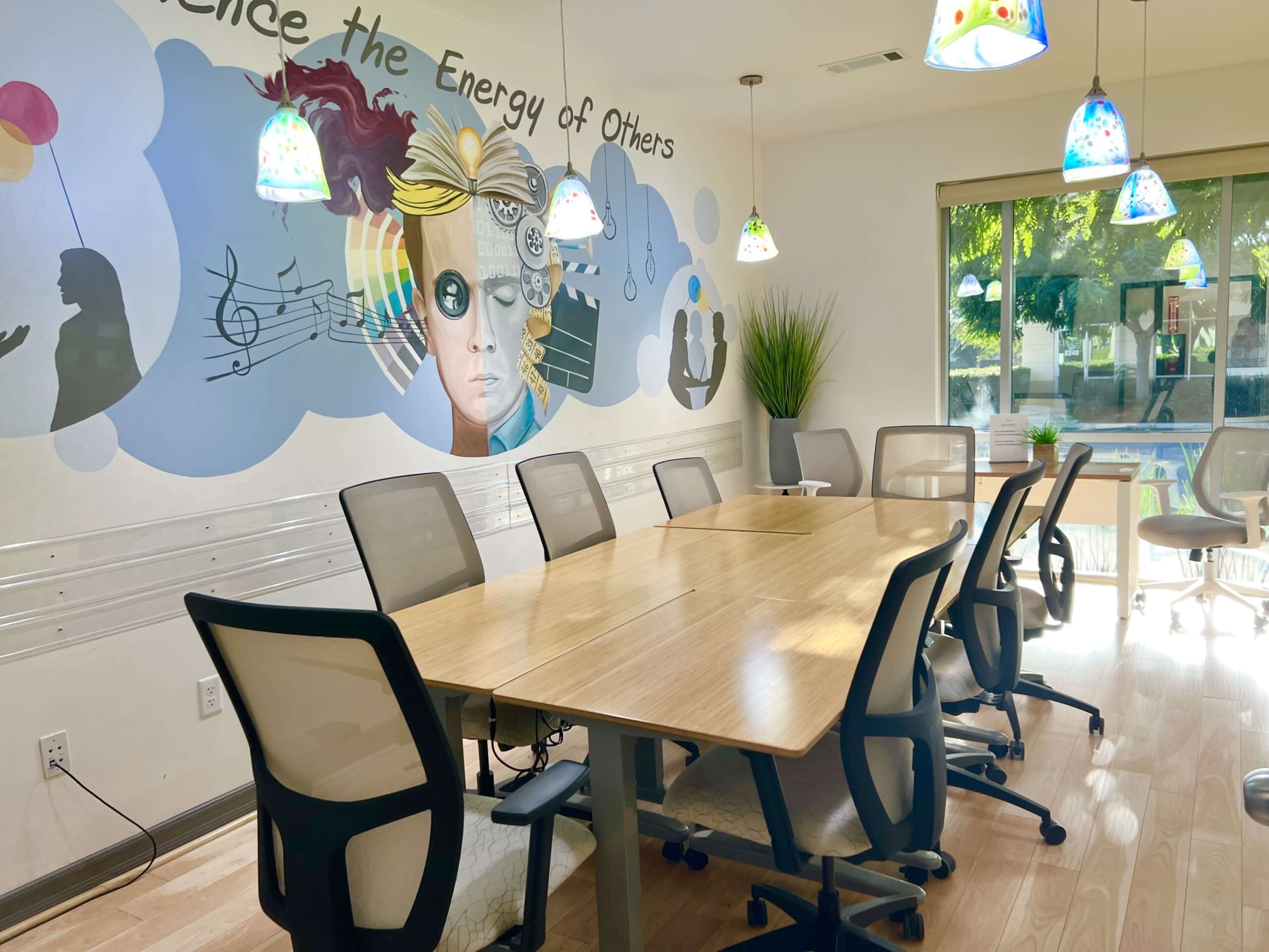 The image shows a modern conference room featuring a long wooden table surrounded by office chairs, with a mural on one wall and decorative light fixtures hanging from the ceiling.