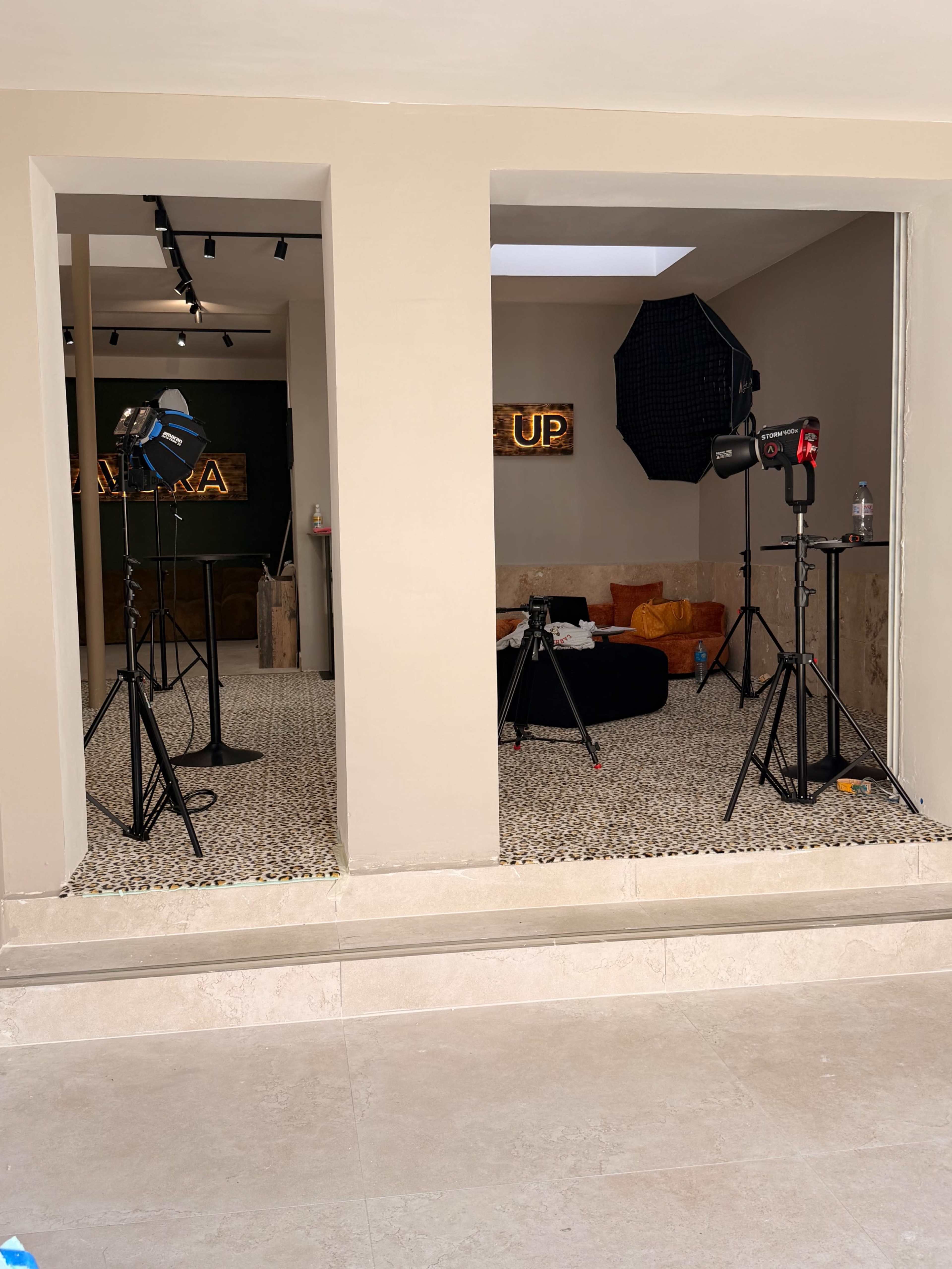 The image shows a studio setup with lighting equipment and a makeup area, featuring a decorative wall and a colorful couch.