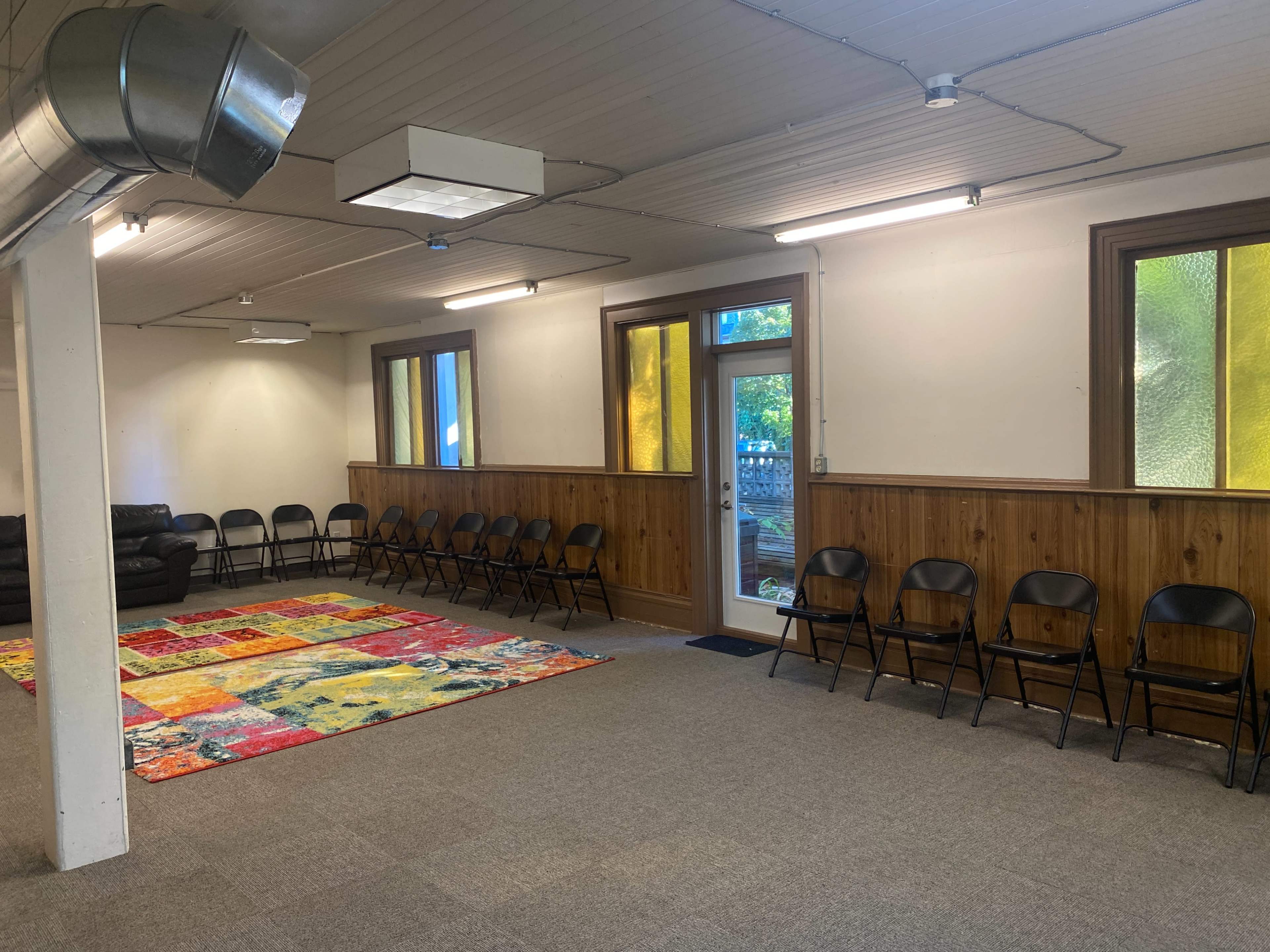 A large room with a carpet in the center, surrounded by rows of black folding chairs and natural light coming through windows.