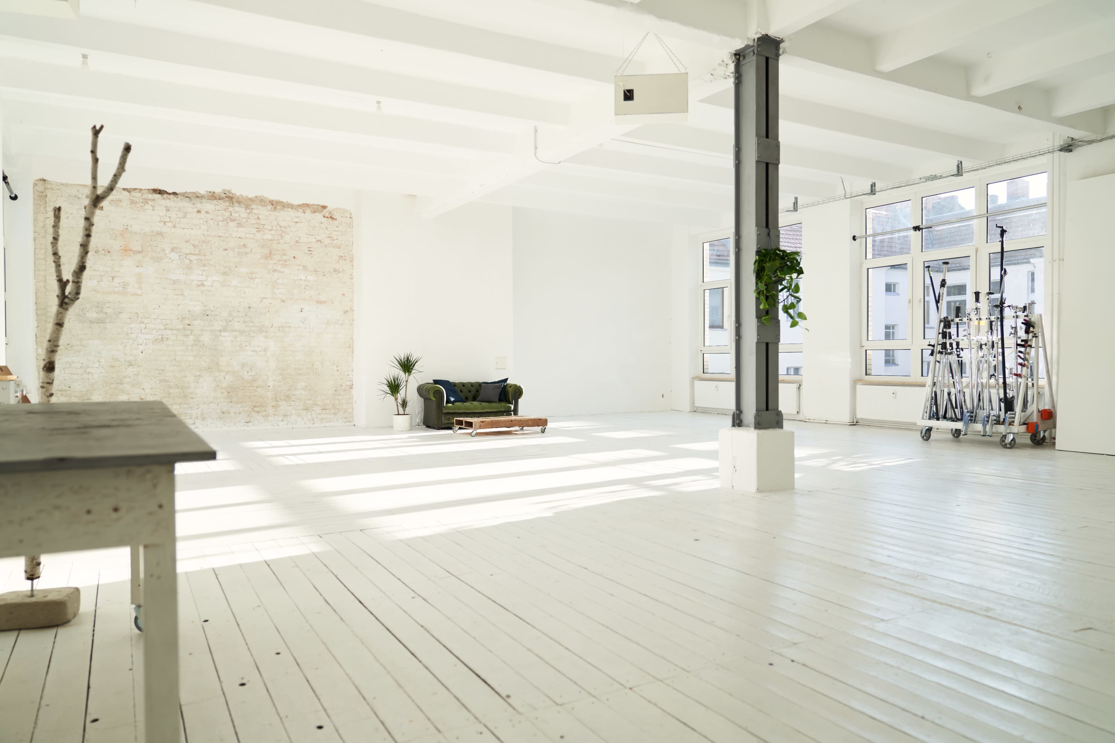 A spacious, minimalist room with white wooden floors, large windows, and a green sofa against a brick wall.