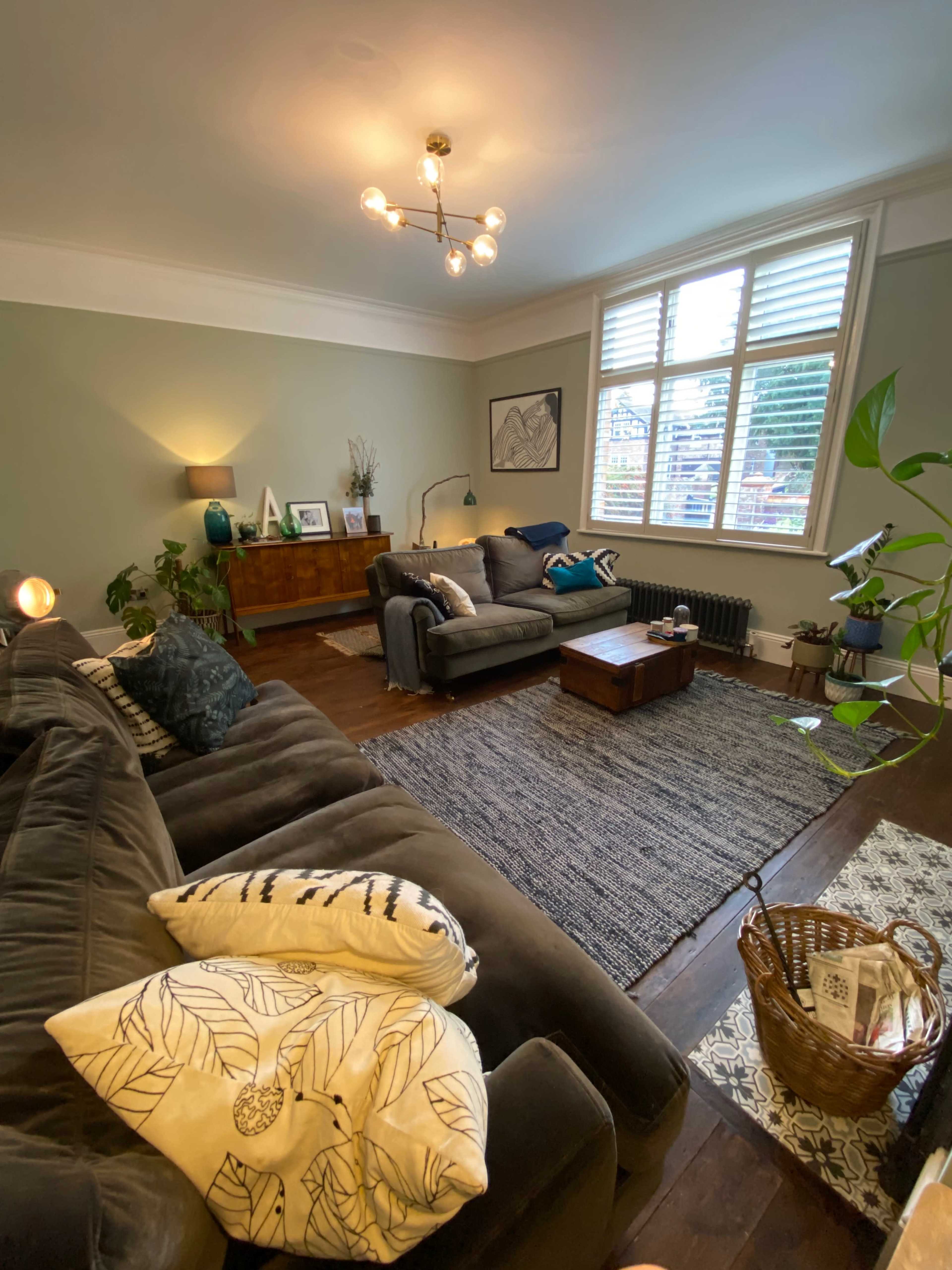 The living room features two sofas, a coffee table, and a large window with shutters, surrounded by decorative plants and a rug.