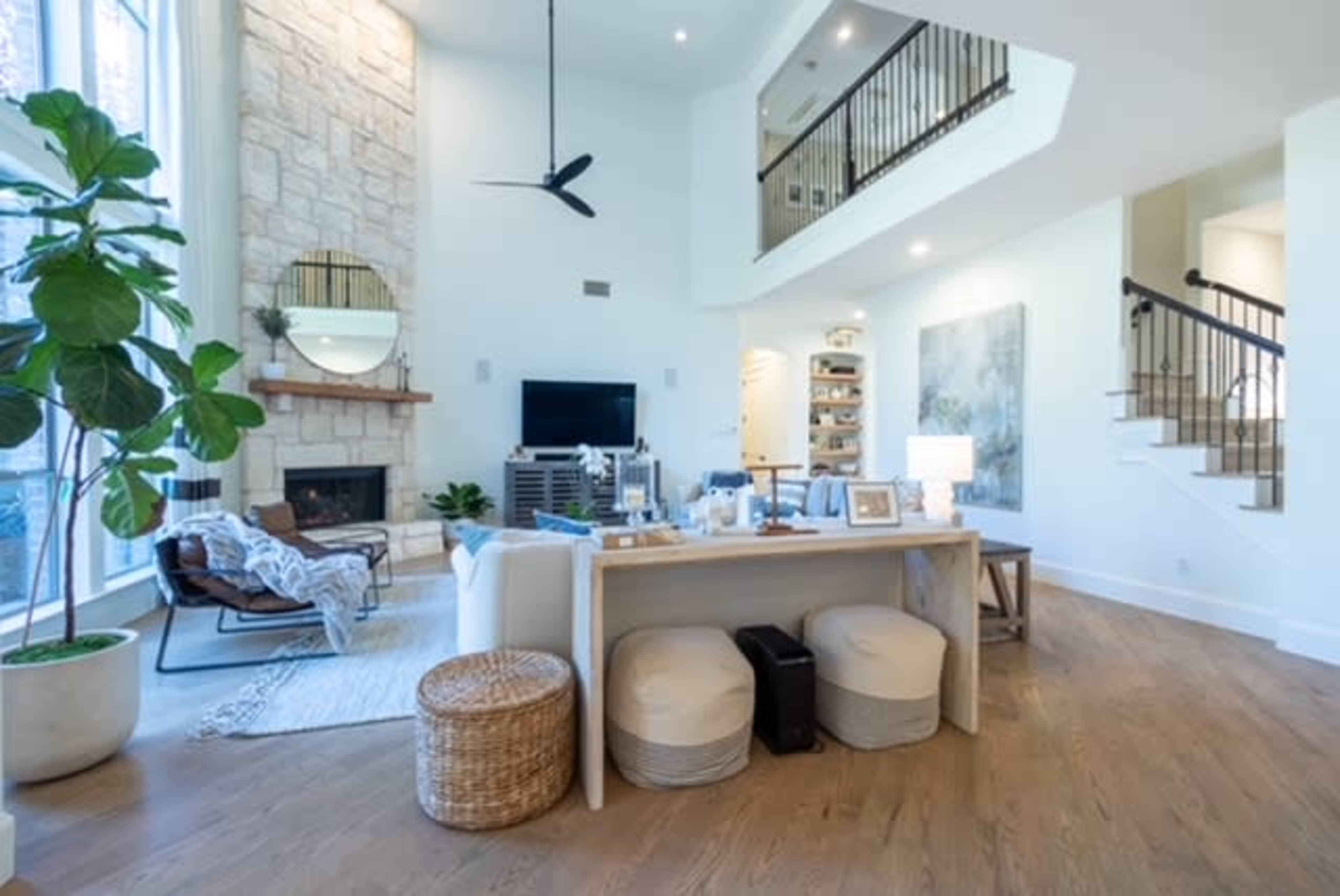 A modern living room with a stone fireplace, a large window, and an open staircase, featuring a wooden table and various seating options.