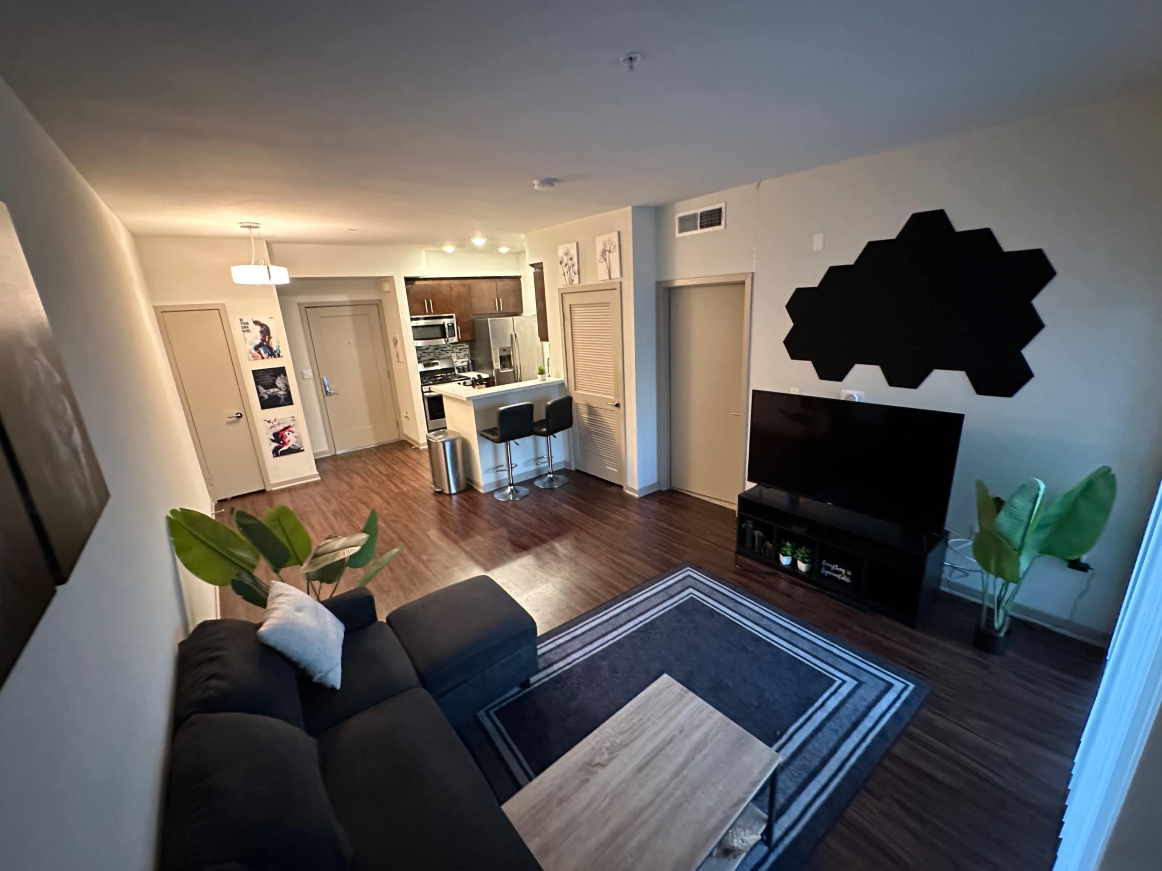 A modern living room with a dark gray sectional sofa, a coffee table, and a TV stand, leading into an open kitchen area with bar stools.