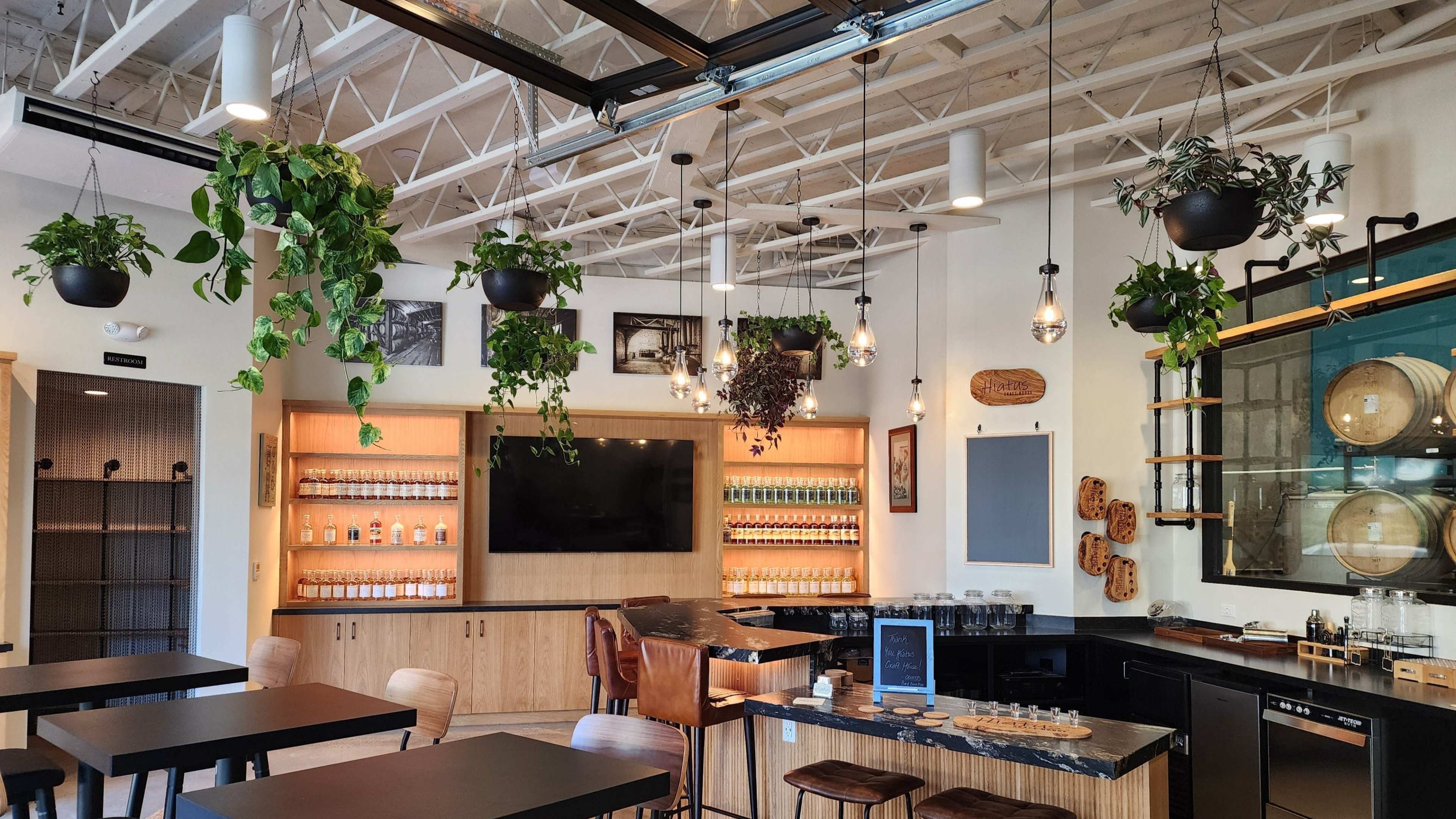 The image shows a modern café interior with wood and glass elements, featuring hanging plants, a large television, shelves of beverages, and multiple seating areas.