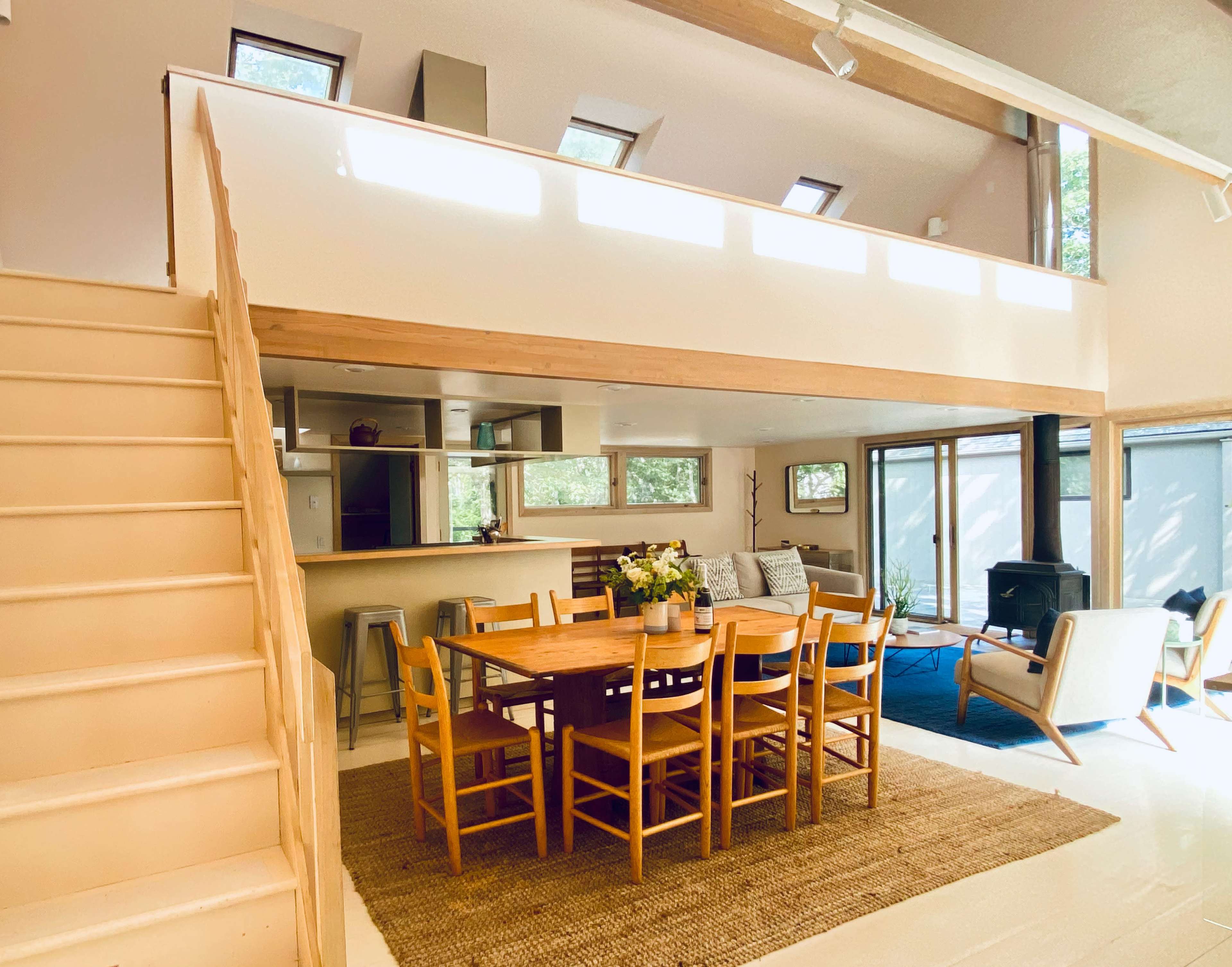 A modern, open-concept living and dining area with a wooden dining table surrounded by chairs, a staircase leading to a loft, and large windows providing natural light.