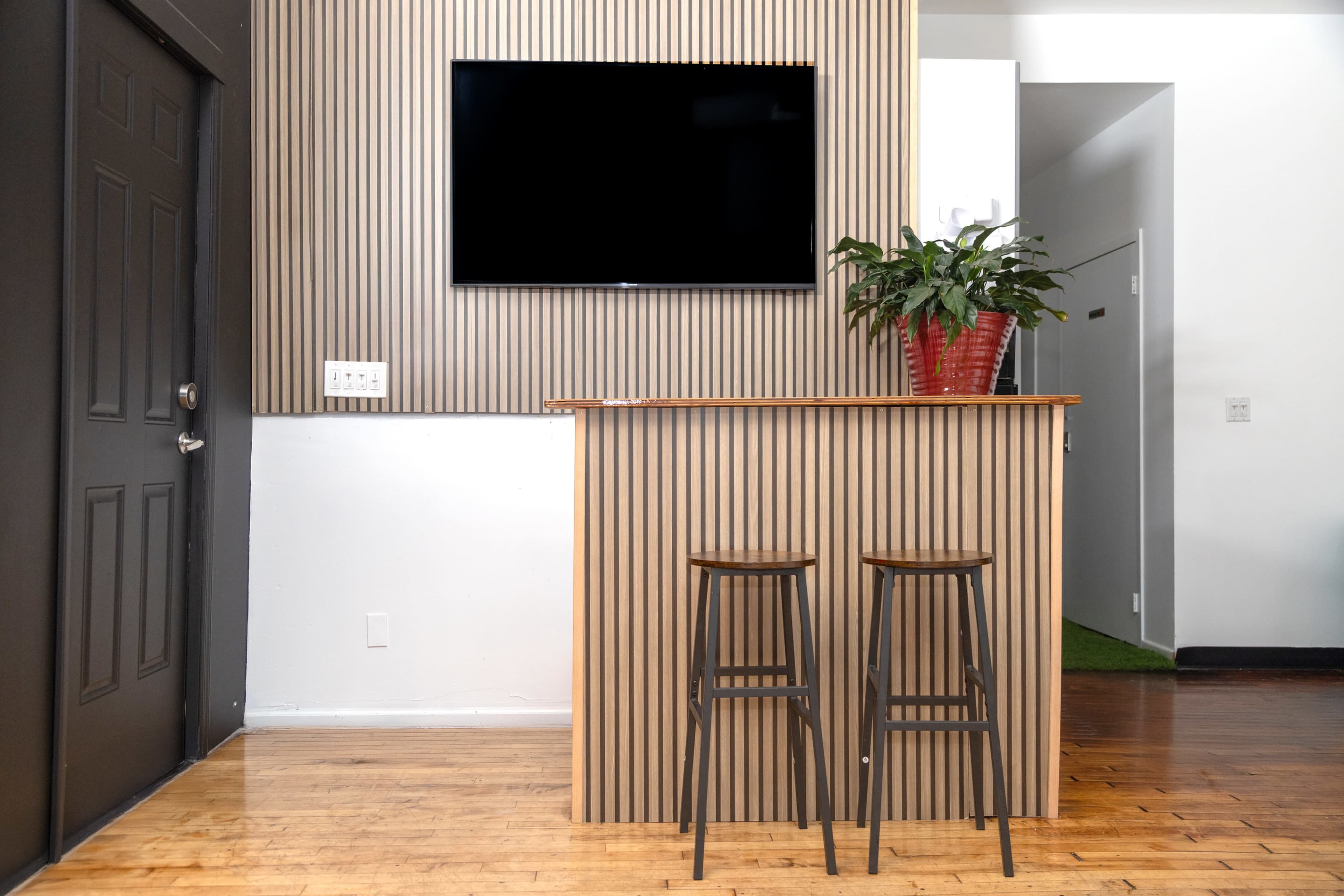 A minimalist interior features a wooden bar with two stools, a wall-mounted television, and a potted plant against a striped wall.