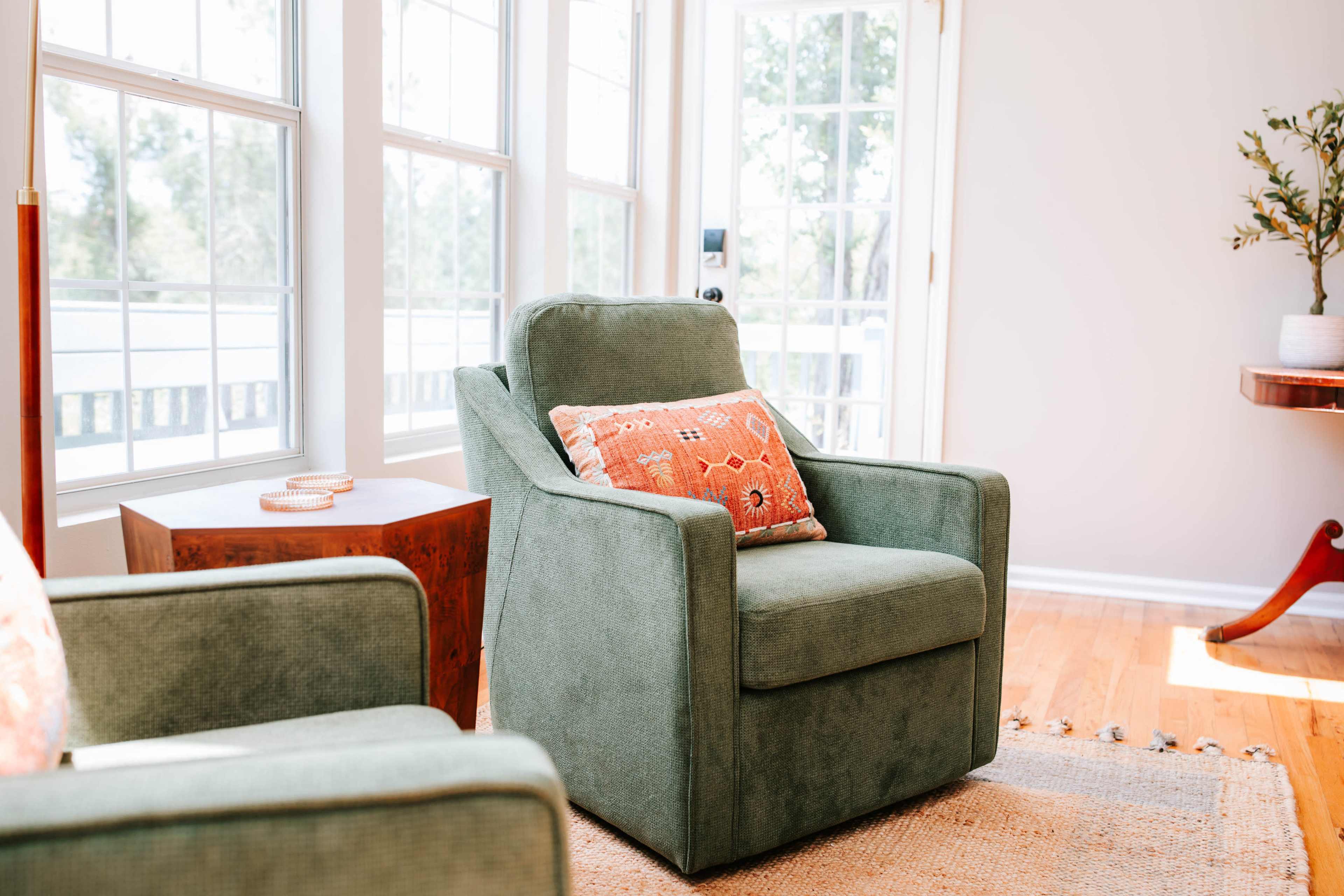 A green upholstered armchair with an orange patterned cushion is positioned next to a wooden side table in a sunlit room with large windows.