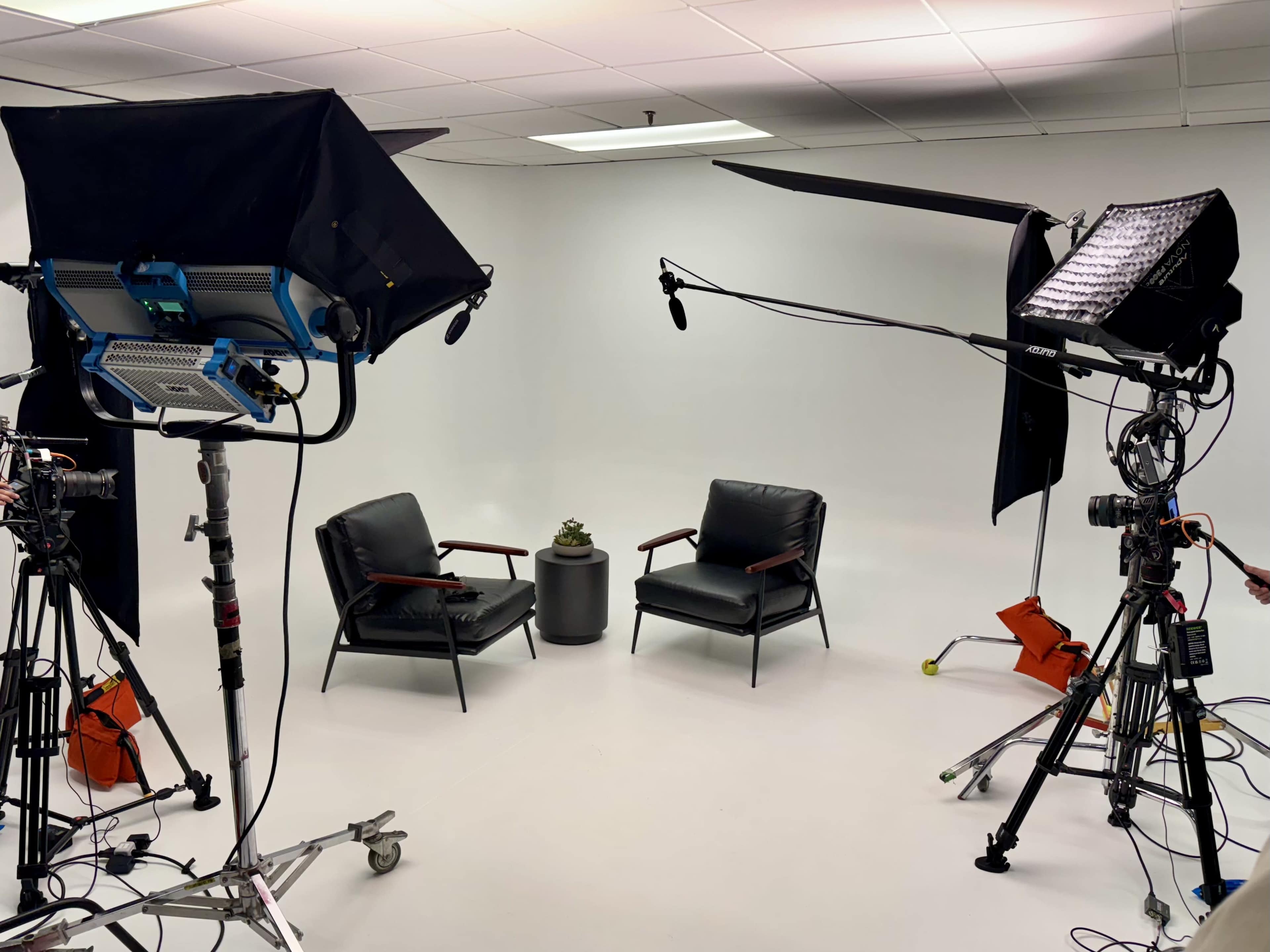 The image shows a recording studio setup with two black chairs facing each other, a small plant on a table, and various lighting and camera equipment arranged around the scene.