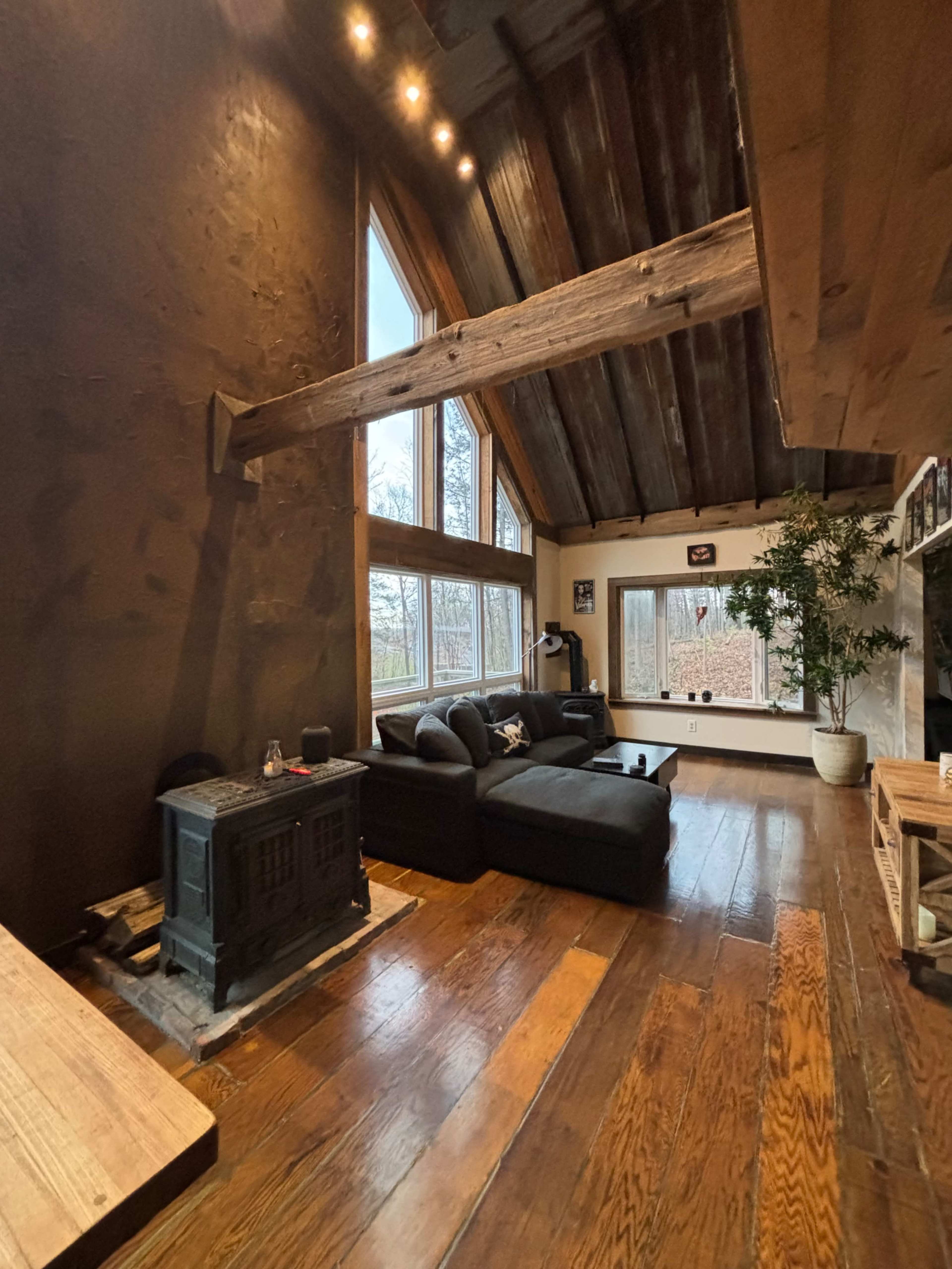 The living room features large windows, a black sofa, a wood stove, and exposed wooden beams.
