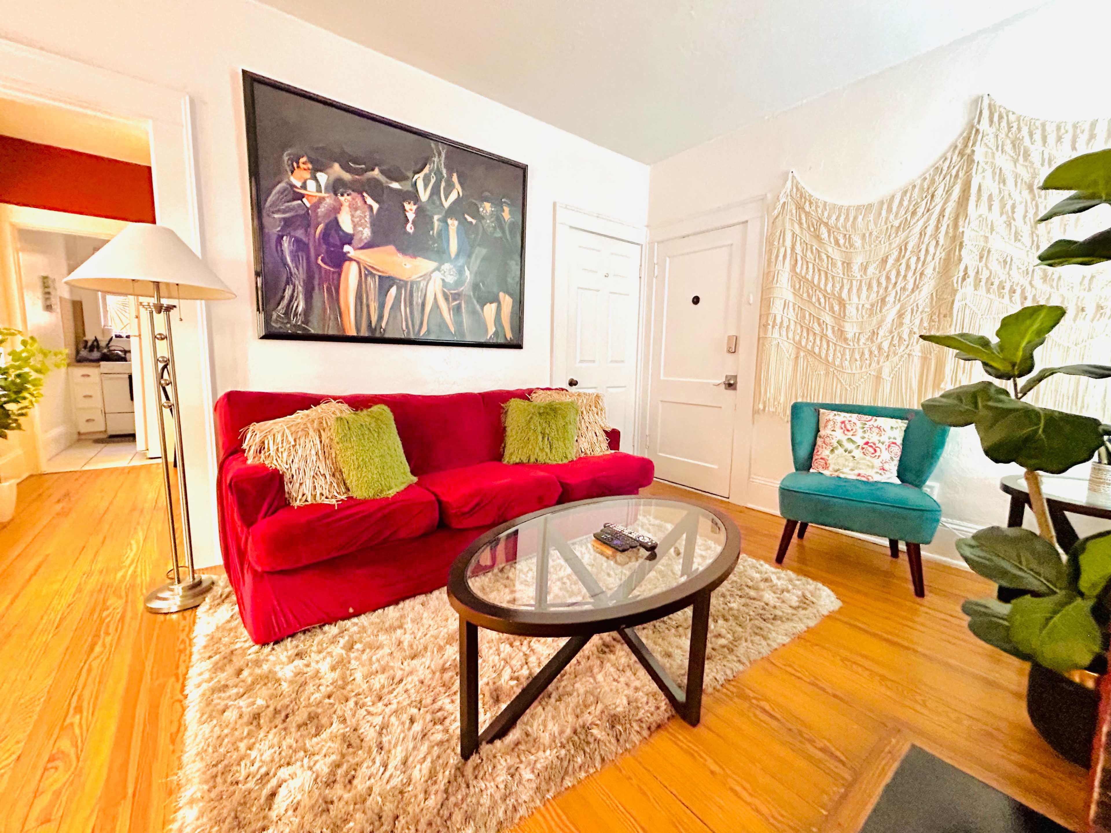 A living room features a red sofa with decorative pillows, a glass coffee table, and a large painting, alongside a teal chair and a textured wall hanging.