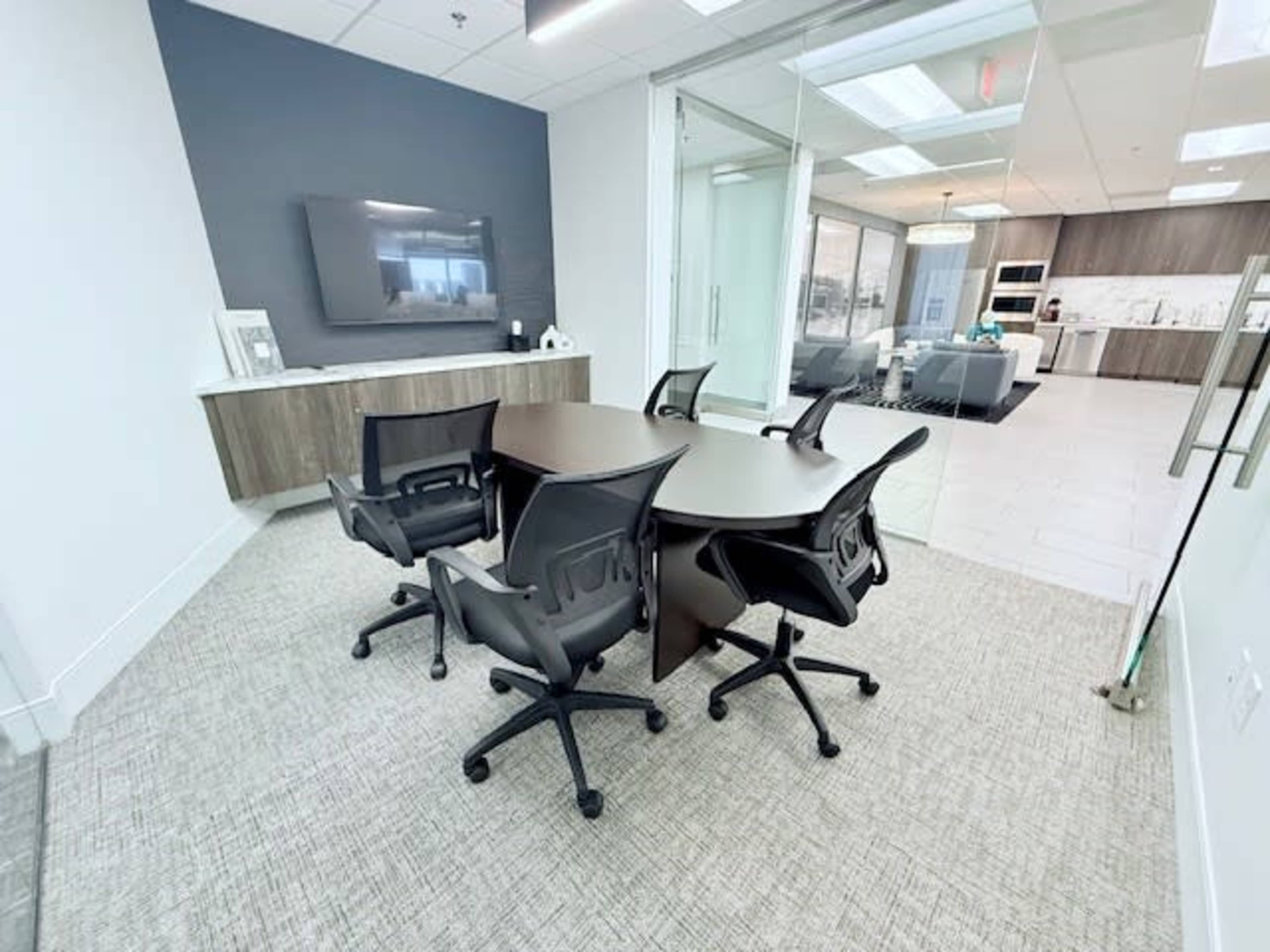 The image shows a modern conference room featuring a round table with four swivel chairs, a large wall-mounted television, and a glass wall that leads to a kitchen area.