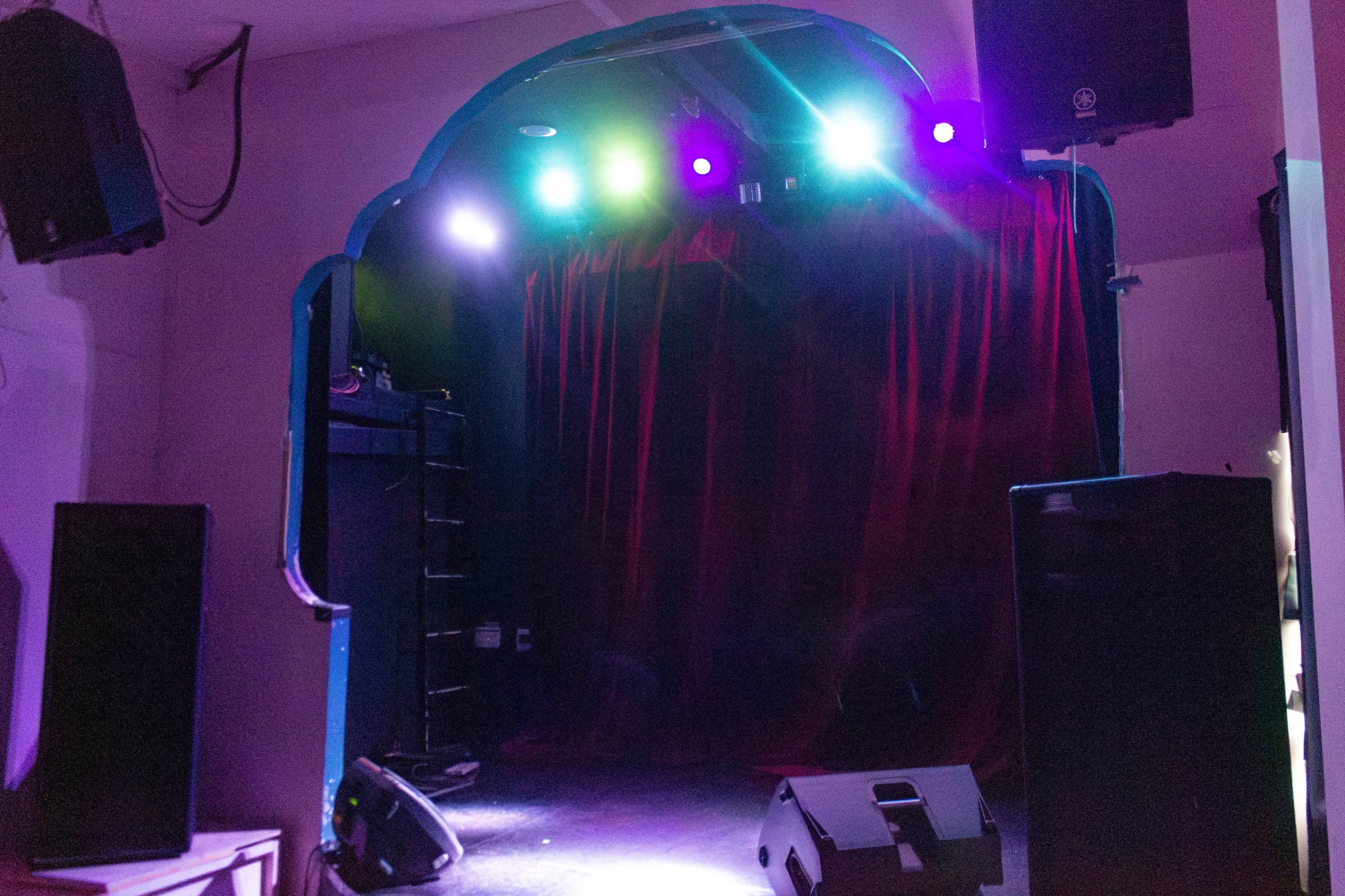 A stage with a red curtain, flanked by large speakers, and illuminated by colorful lights.