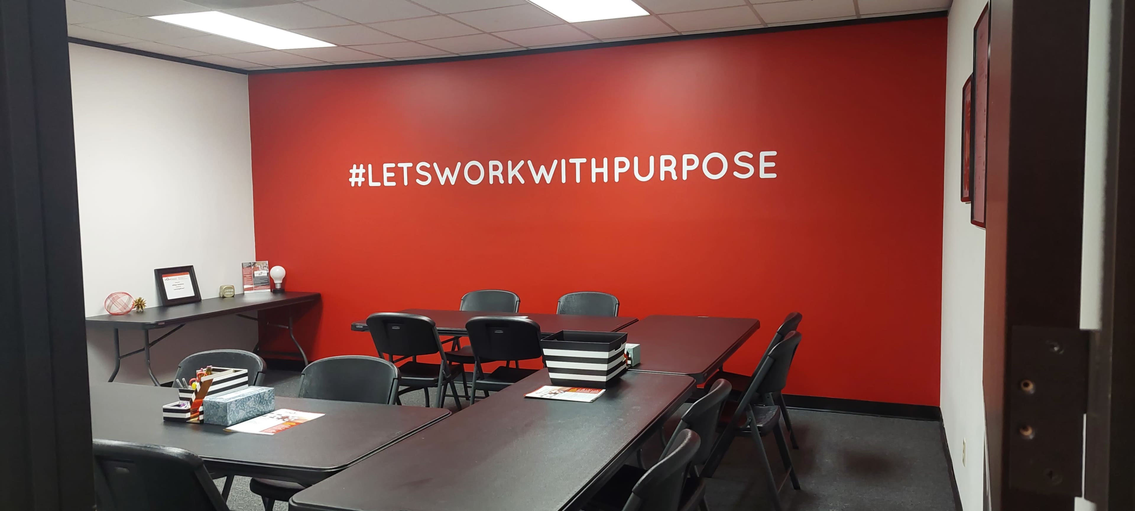 The image shows a meeting room with several black chairs and tables arranged under a vibrant red wall featuring the phrase "#LETSWORKWITHPURPOSE."