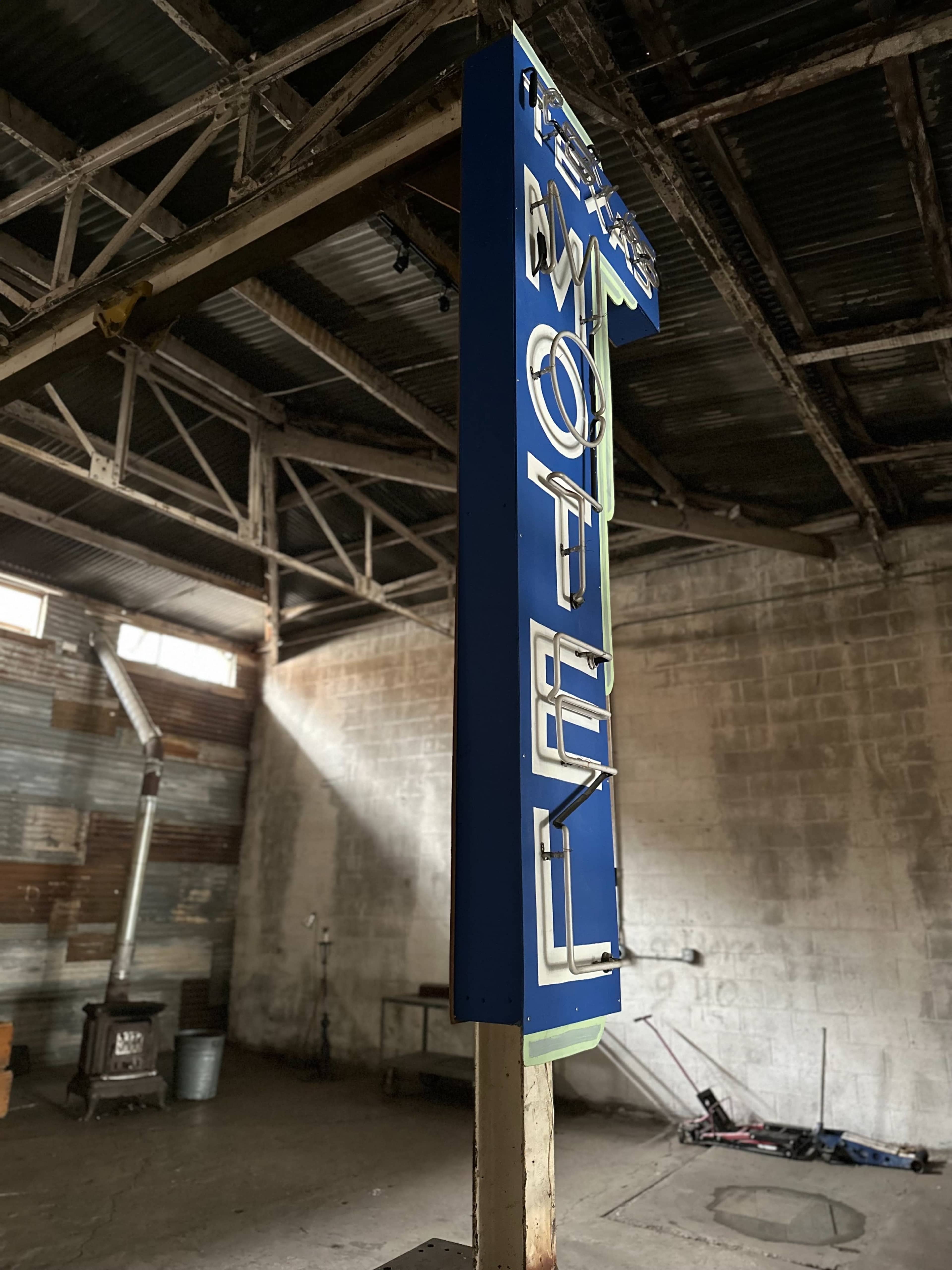 A vintage motel sign standing upright in an industrial, dimly lit indoor space with exposed beams and a concrete wall.