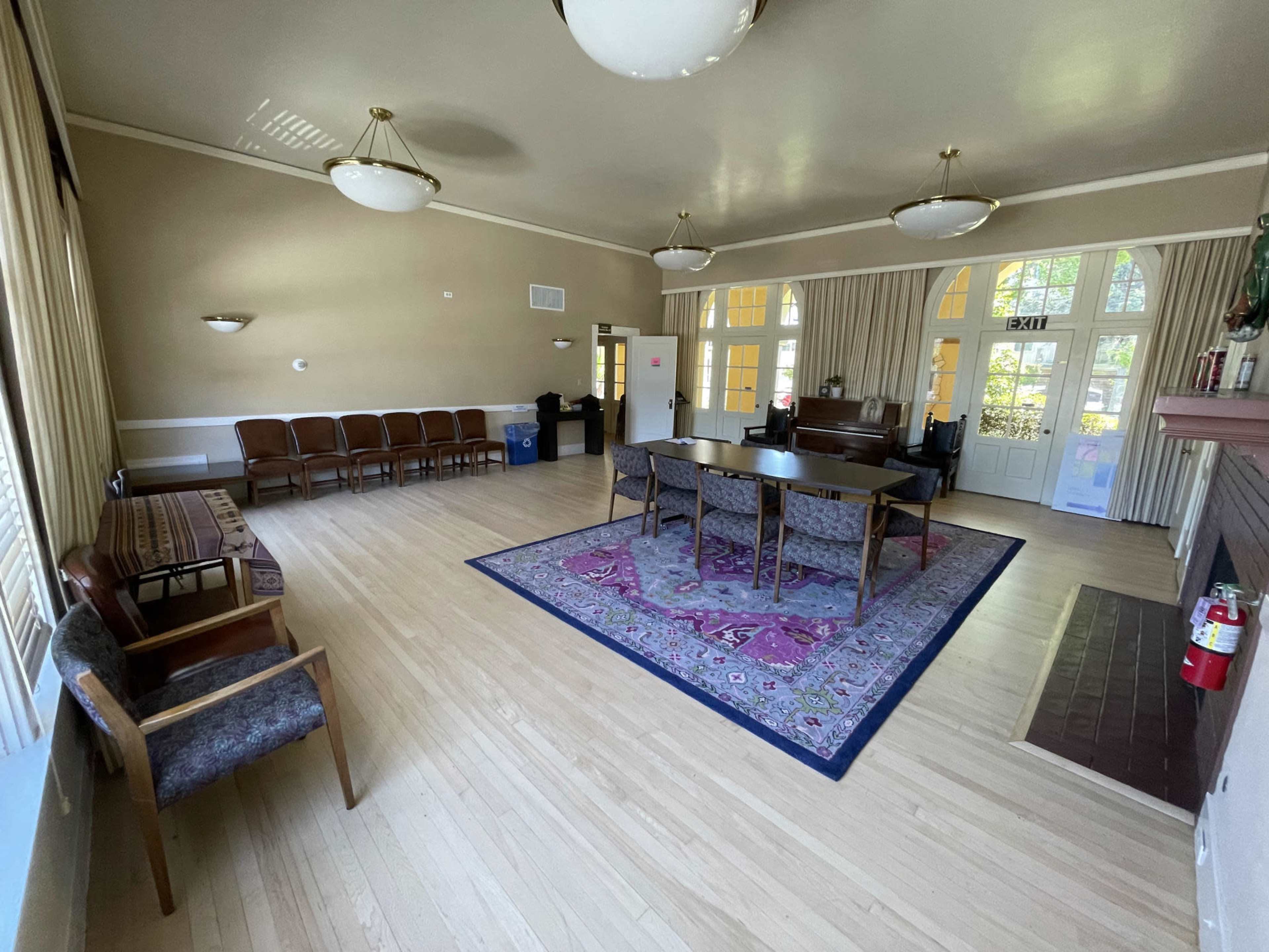 The image shows a spacious room with wooden floors, a table and chairs in the center, and a piano against one wall, illuminated by overhead lights.