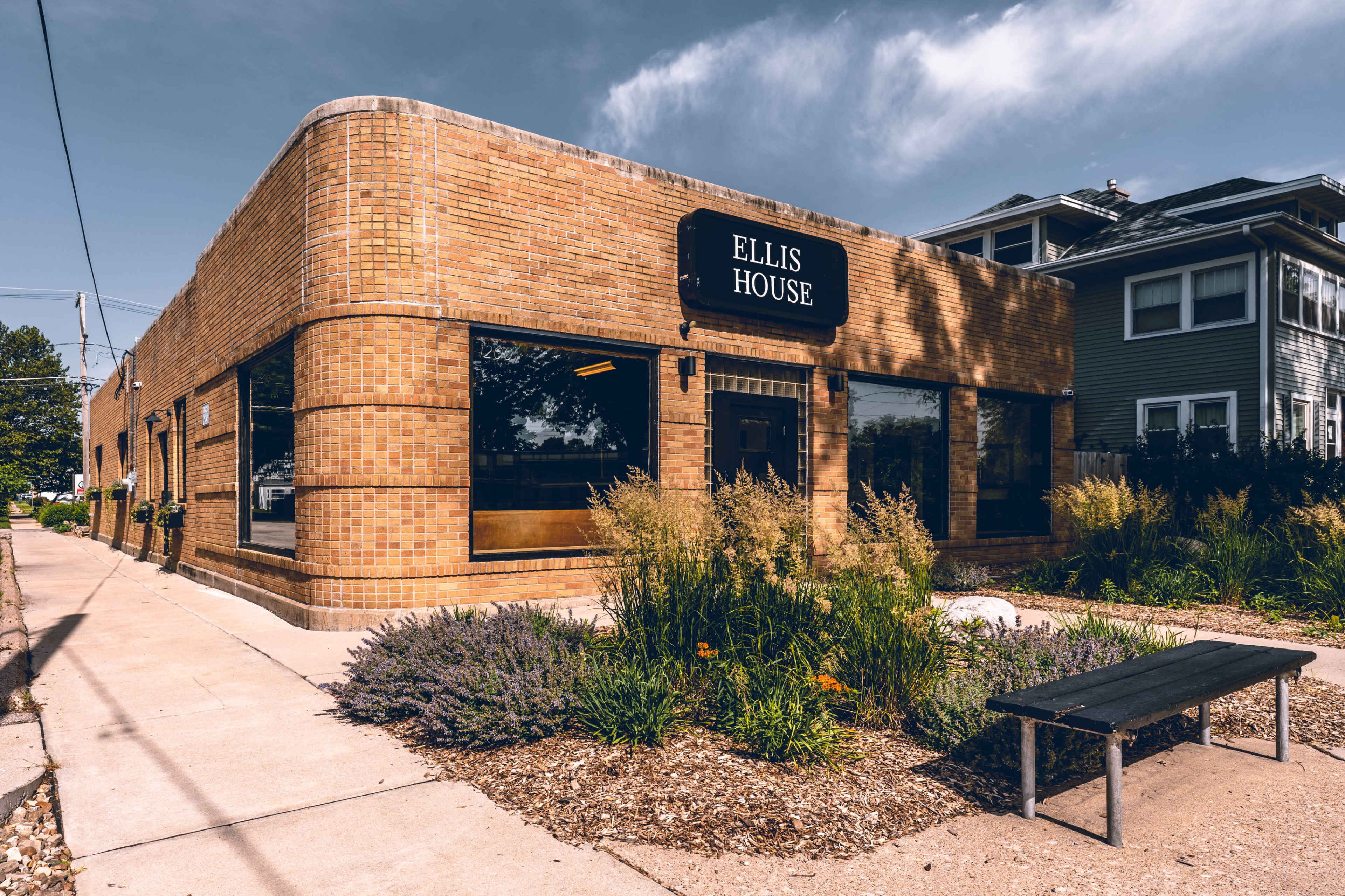 The image shows a brick building labeled "Ellis House" with large windows, surrounded by landscaping and a bench on the sidewalk.