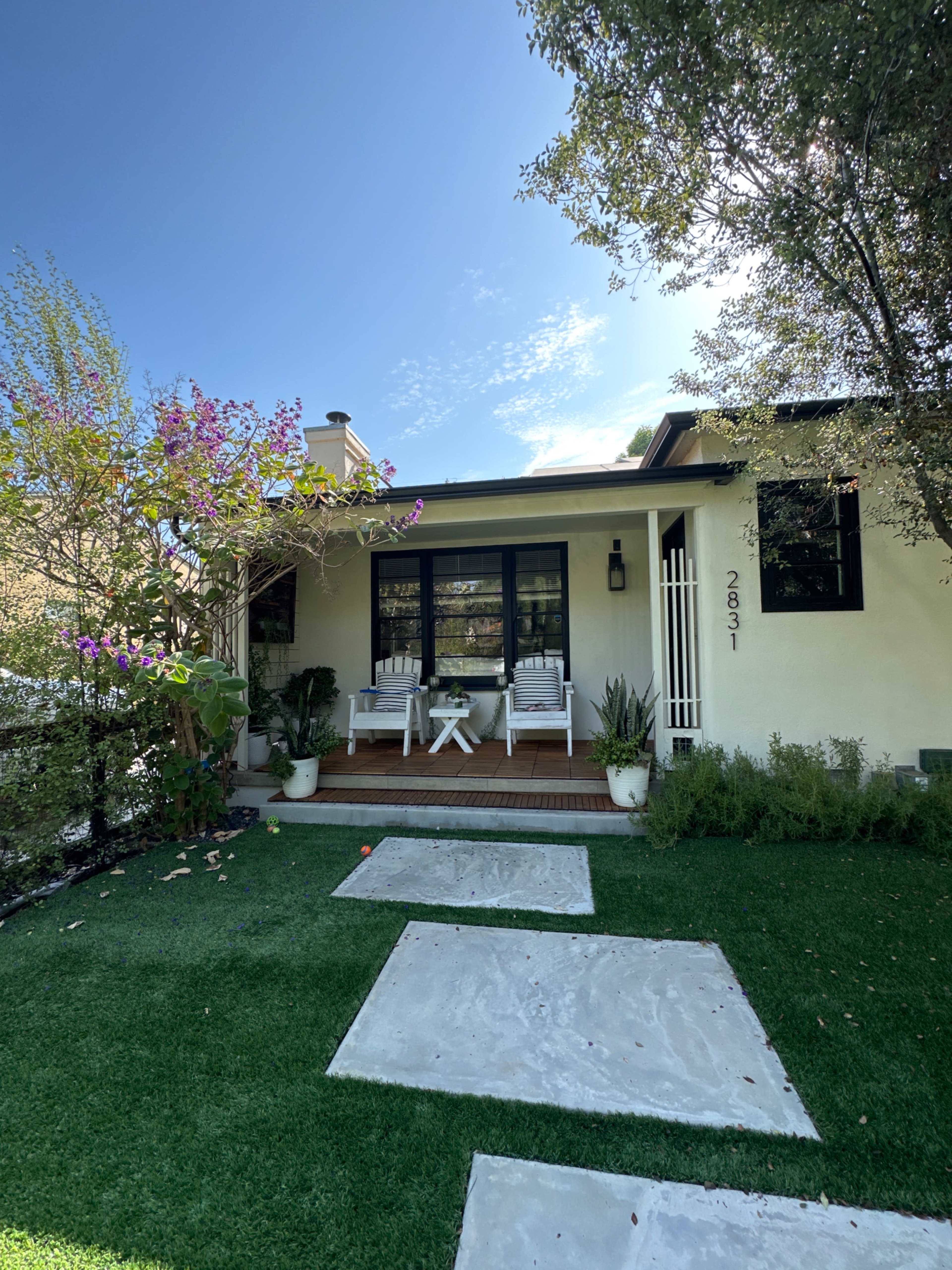 A residential house with a front porch featuring two white chairs, surrounded by green grass and flowering plants.