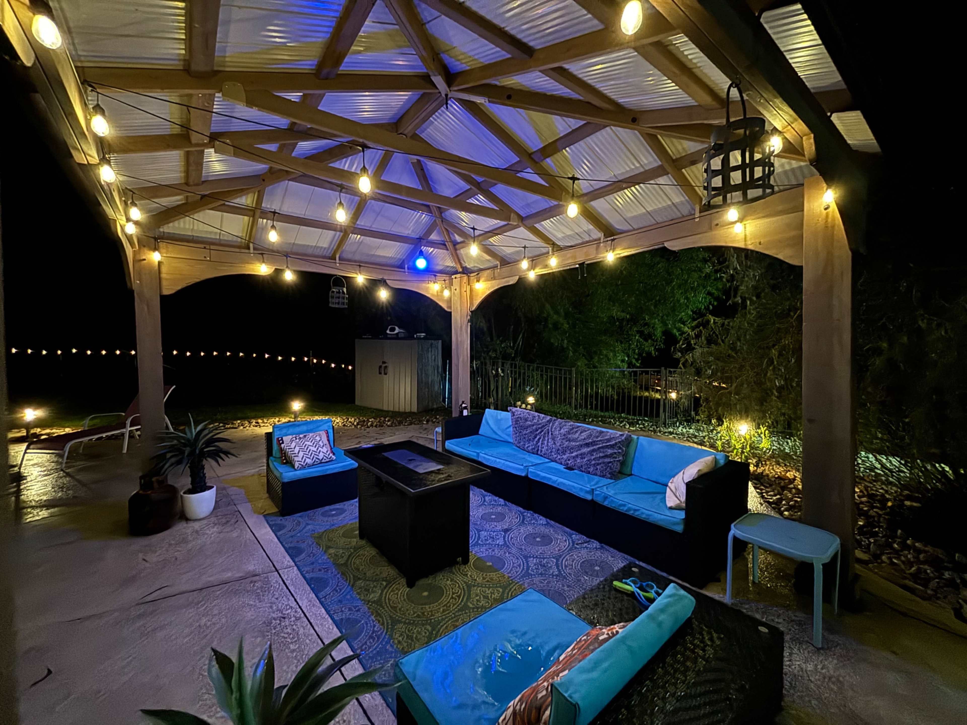A covered outdoor seating area features a sectional couch, a coffee table, and string lights illuminating the space at night.