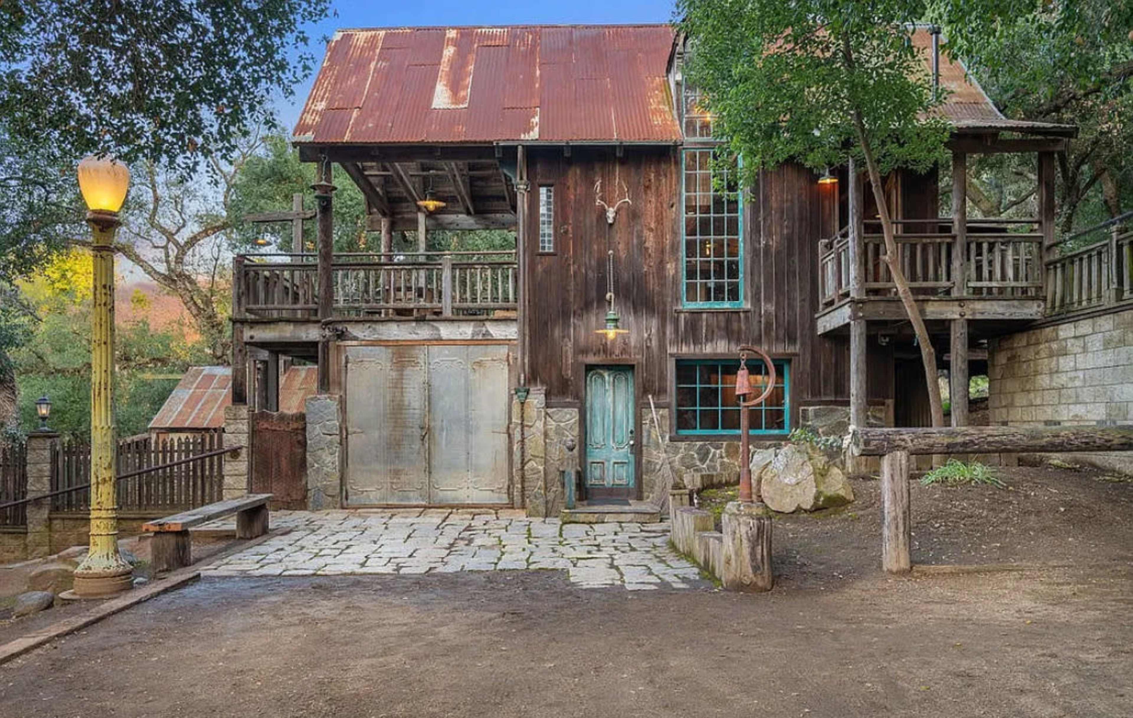 The image shows a rustic wooden building with a corrugated metal roof, featuring multiple balconies and a stone path leading to its entrance.