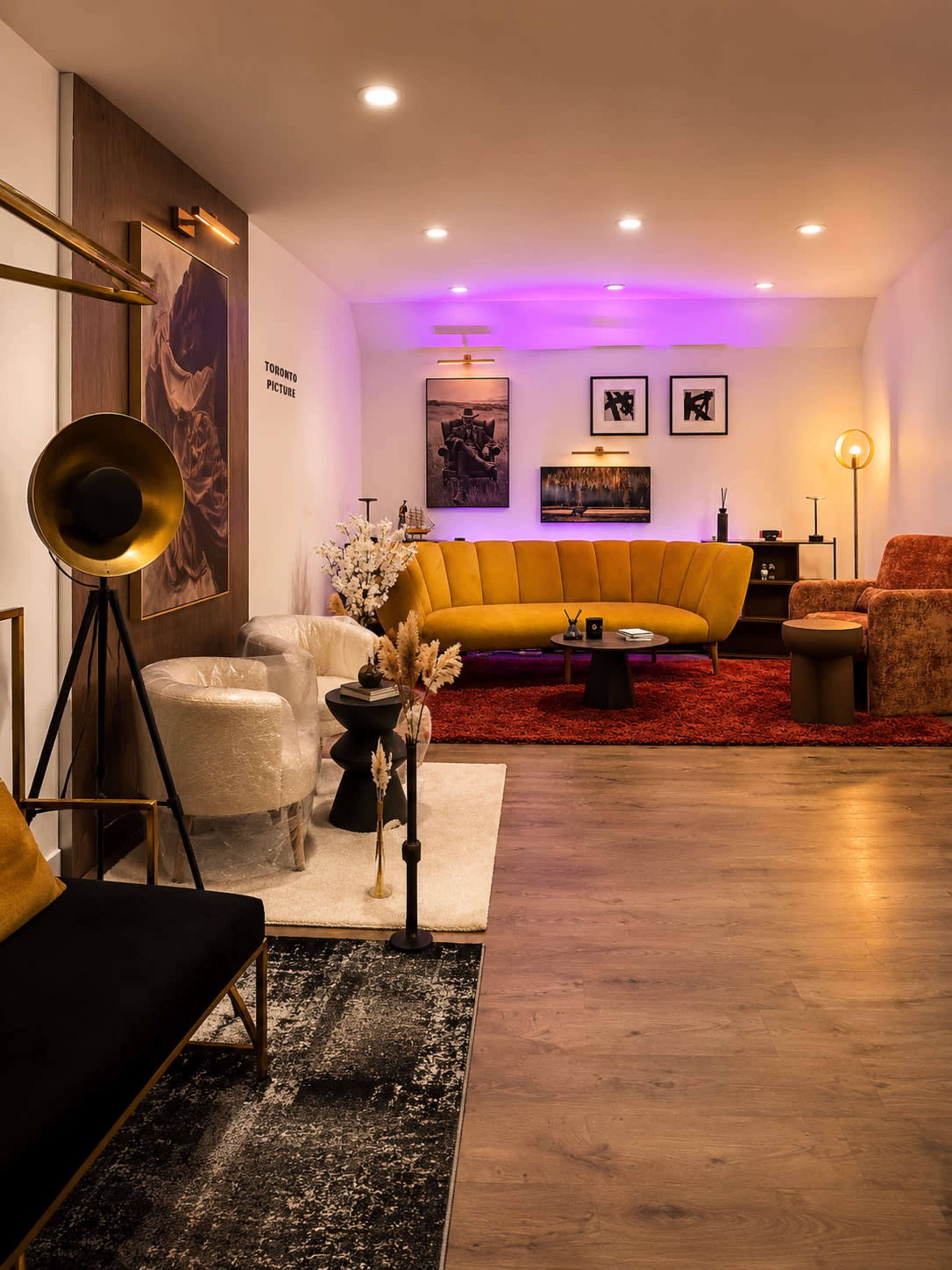The image shows a stylish interior living space featuring a curved yellow sofa, various artworks on the walls, modern lighting fixtures, and a mix of seating arrangements.