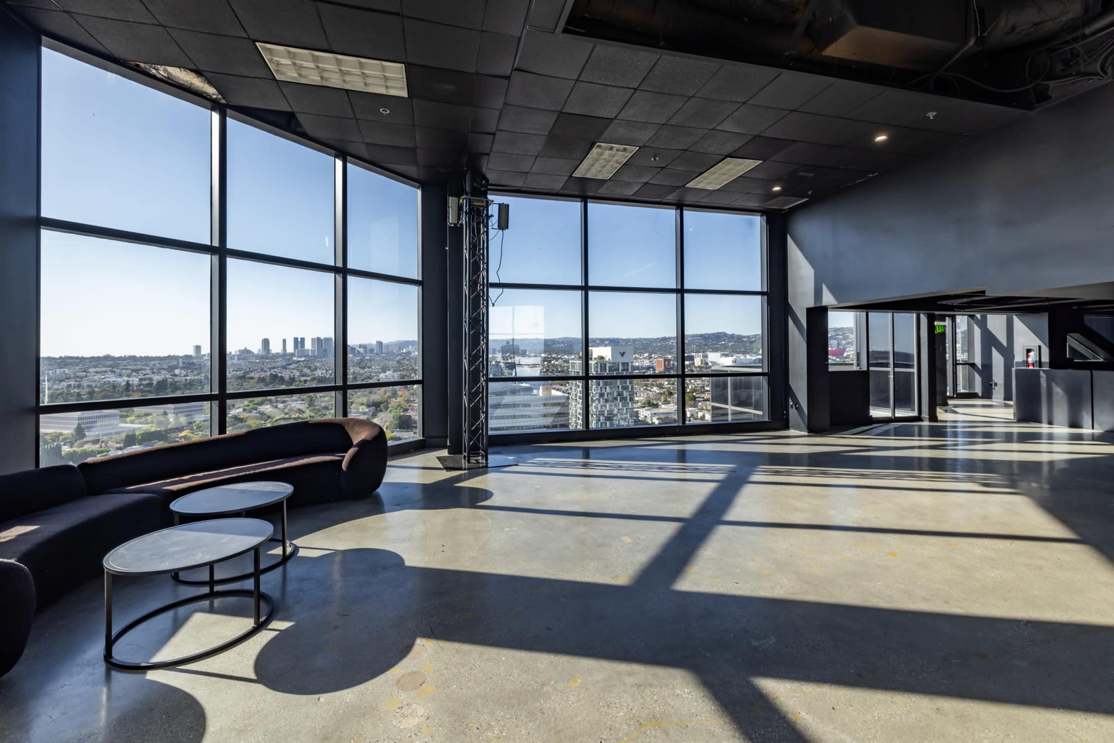 The image shows a modern interior space with large windows offering a view of the city skyline, featuring a curved couch and round tables on polished concrete flooring.