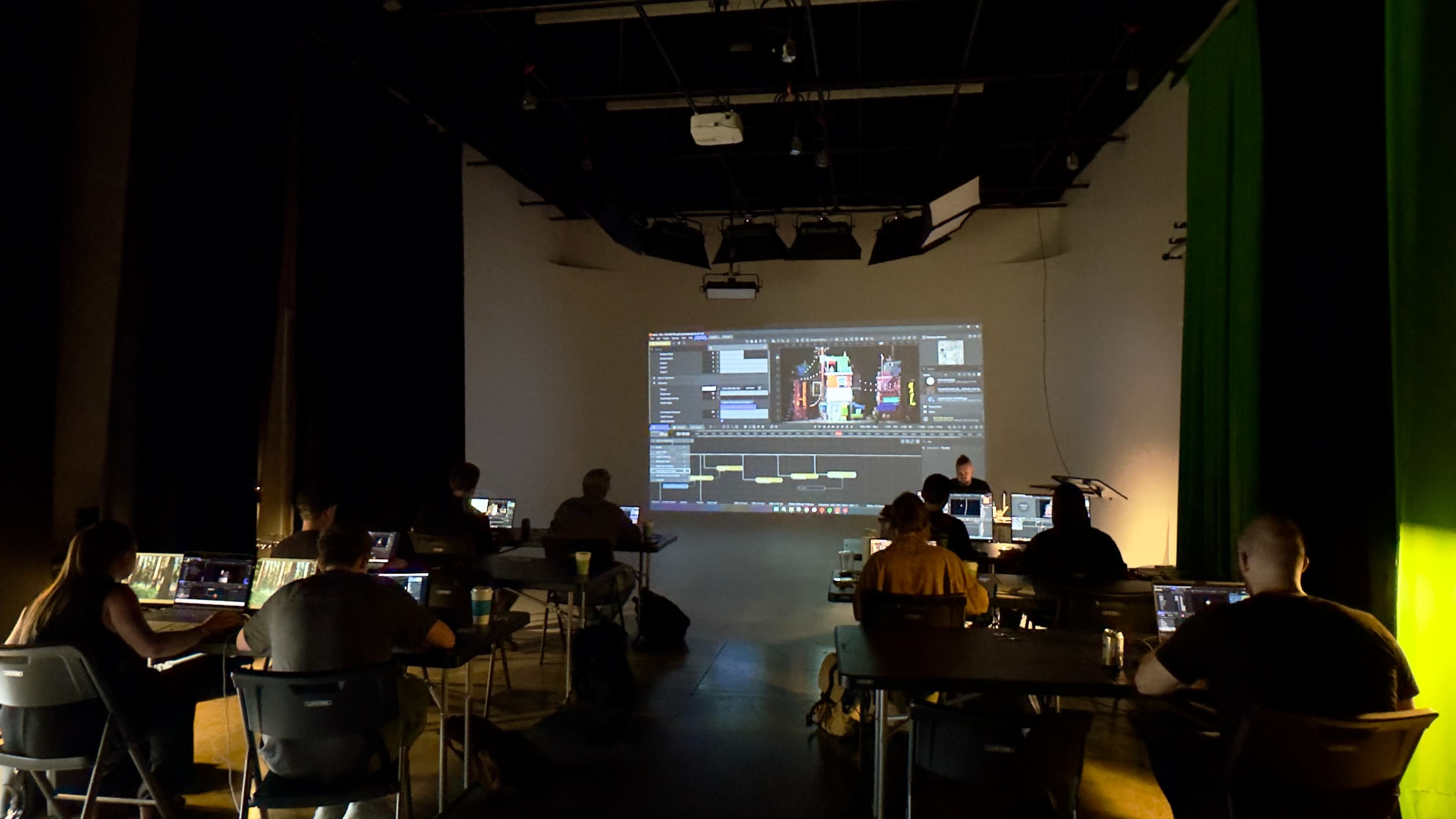 A group of people sit at desks in a dimly lit room, focused on their laptops while a large screen displays editing software.