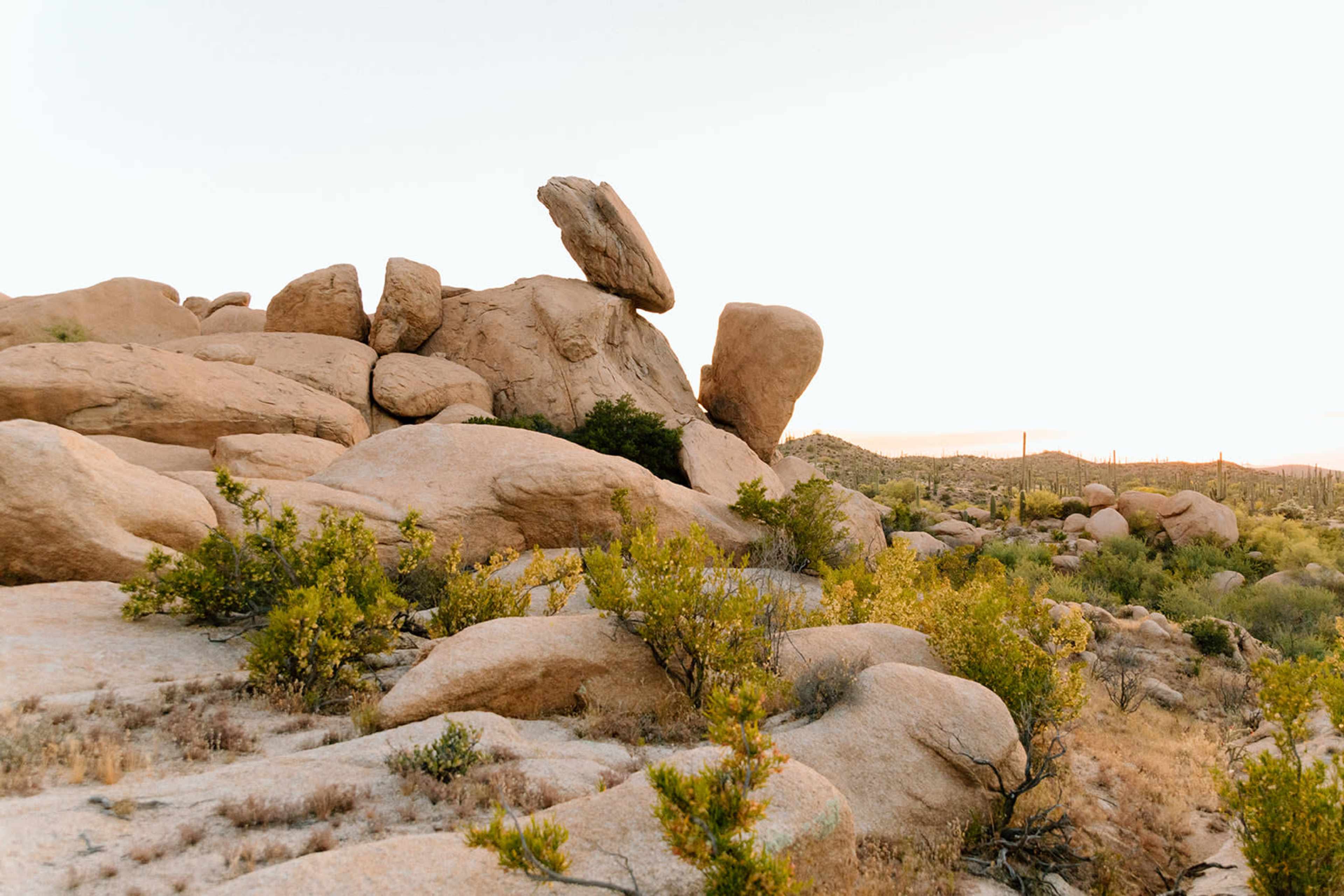 A large boulder precariously balanced atop a cluster of smaller rocks sits amidst a desert landscape with sparse vegetation.