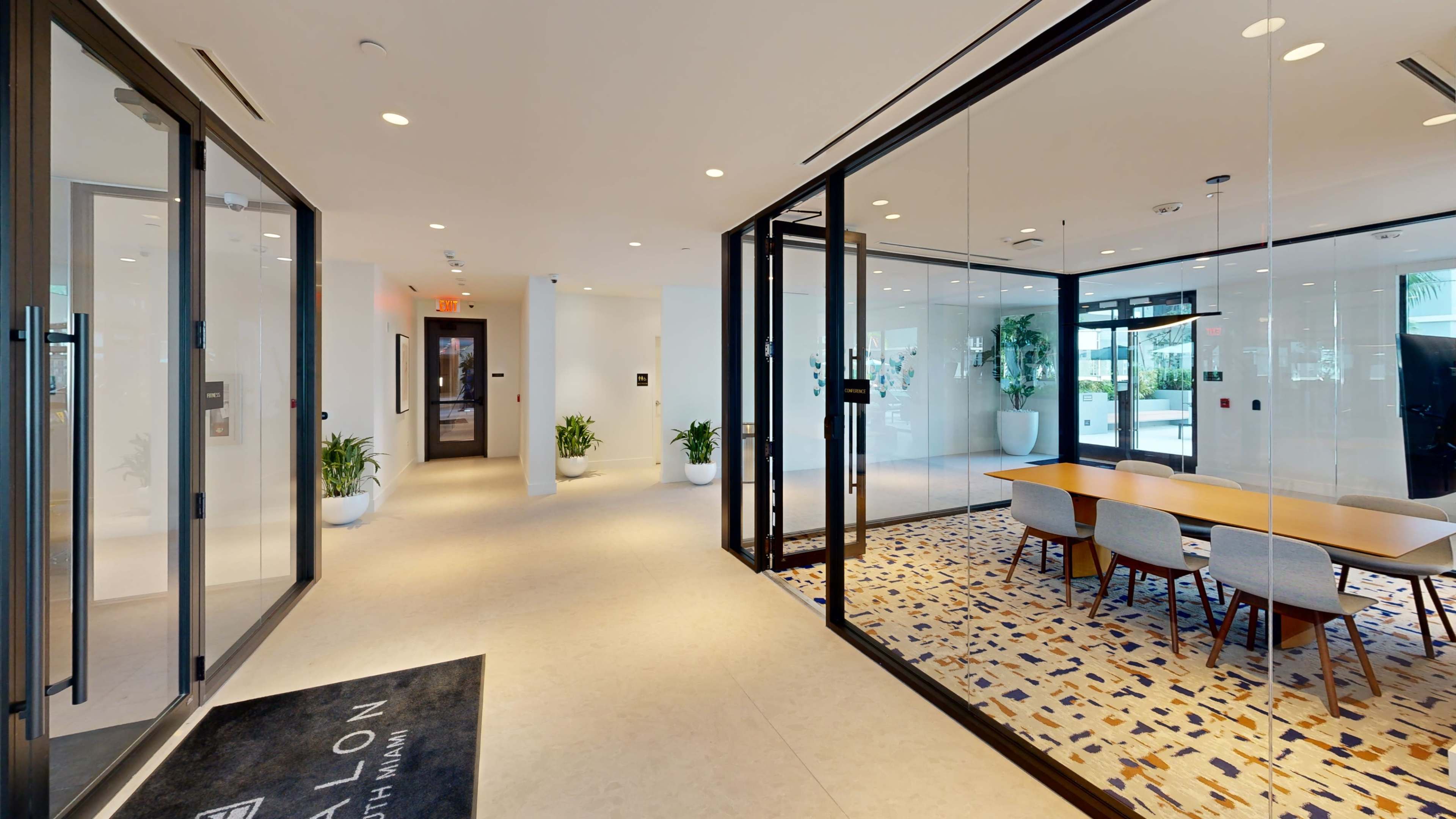The image shows a modern lobby area featuring glass doors, a spacious conference room with a wooden table and chairs, and decorative plants in large pots.
