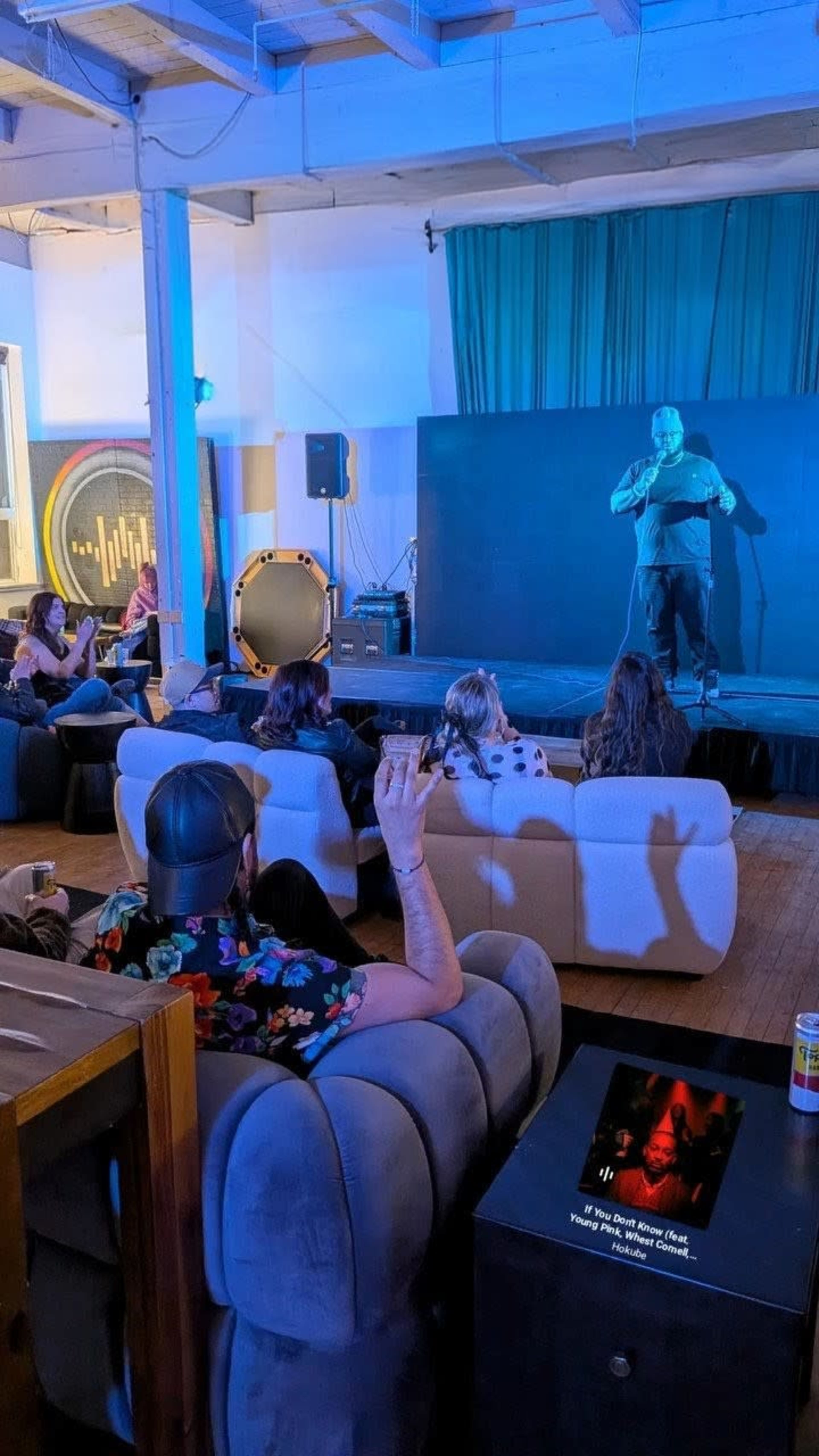 A comedian performs on stage in a dimly lit venue while an audience sits on couches, some engaged and others watching attentively.
