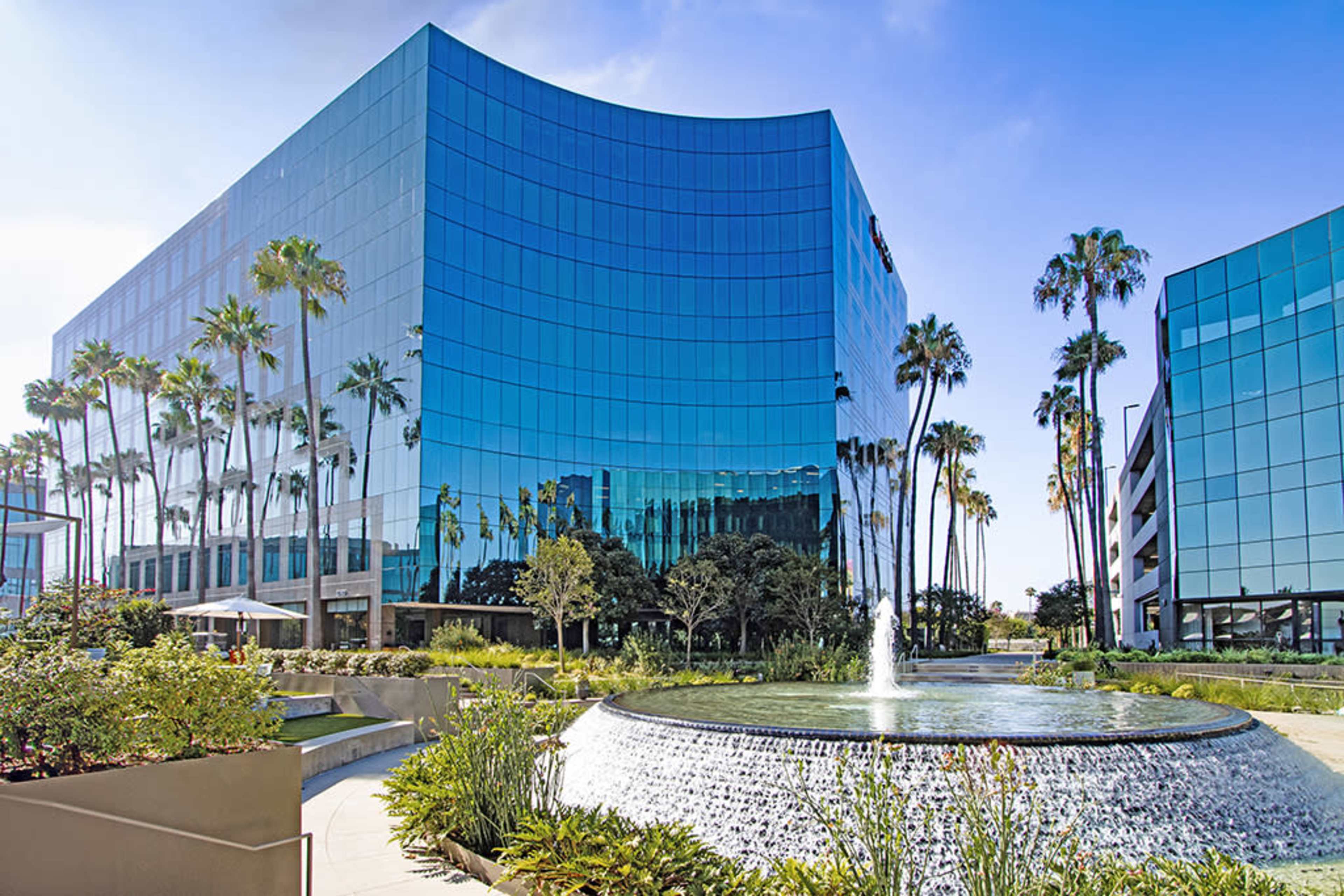 The image shows a modern glass office building reflecting palm trees and a fountain in the foreground.