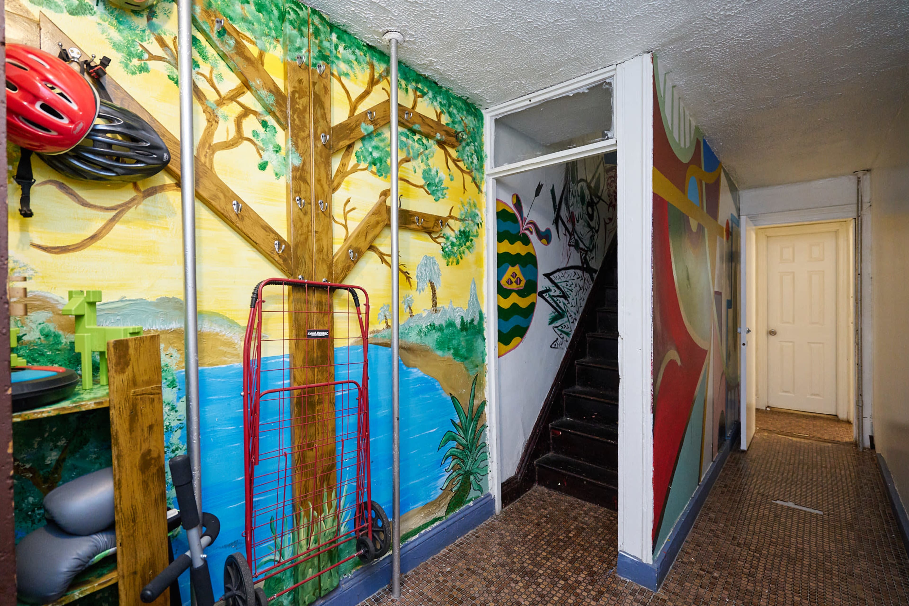 The image depicts a hallway featuring a colorful mural of a tree and water, with a bicycle helmet and a cart mounted on the wall beside a staircase.