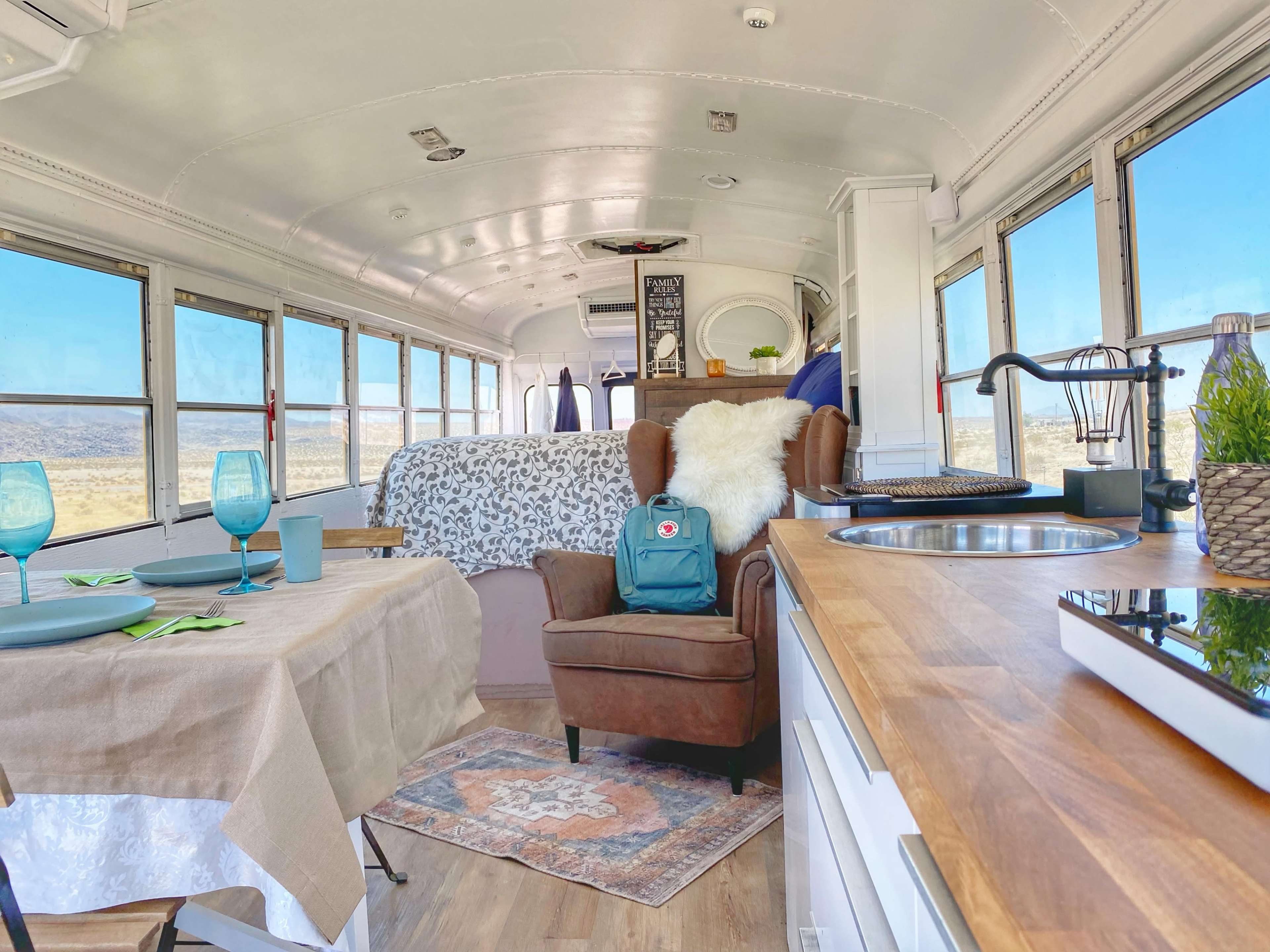 The interior of a converted school bus features a kitchenette, dining area with a table set for two, a comfortable chair, and a bed area lined with large windows that offer views of the landscape outside.